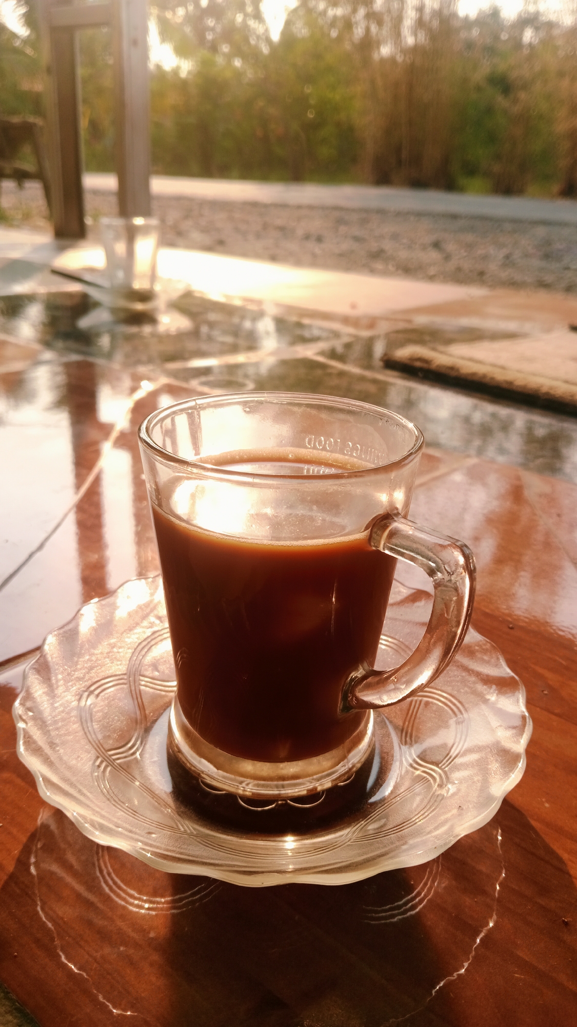 A clear glass mug filled with dark brown liquid, presumably coffee, sits on a clear glass saucer. The mug has a handle and an embossed pattern around its rim. The saucer has a decorative, scalloped edge. Both are placed on a polished wooden surface that reflects light, creating streaks and circular patterns. In the background, the scene is out of focus, showing greenery, trees, and a paved area. Sunlight is casting warm light and shadows. Another empty glass mug is visible further back in the scene, also on a reflective surface. The overall impression is that of a tranquil outdoor setting, possibly a patio or a garden area, in Karang Indah, Indonesia, during the early morning or late afternoon when the sun is low. There are no people or specific actions depicted.