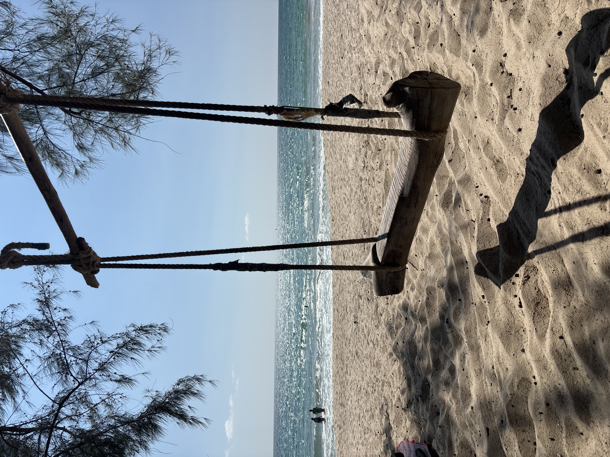 The image shows a wooden swing hanging from a tree overlooking a beach and ocean in Choeng Thale, Thailand. The beach is sandy with shadows cast by the swing and surrounding trees. The ocean stretches to the horizon under a clear blue sky. Two people are standing in the water.
