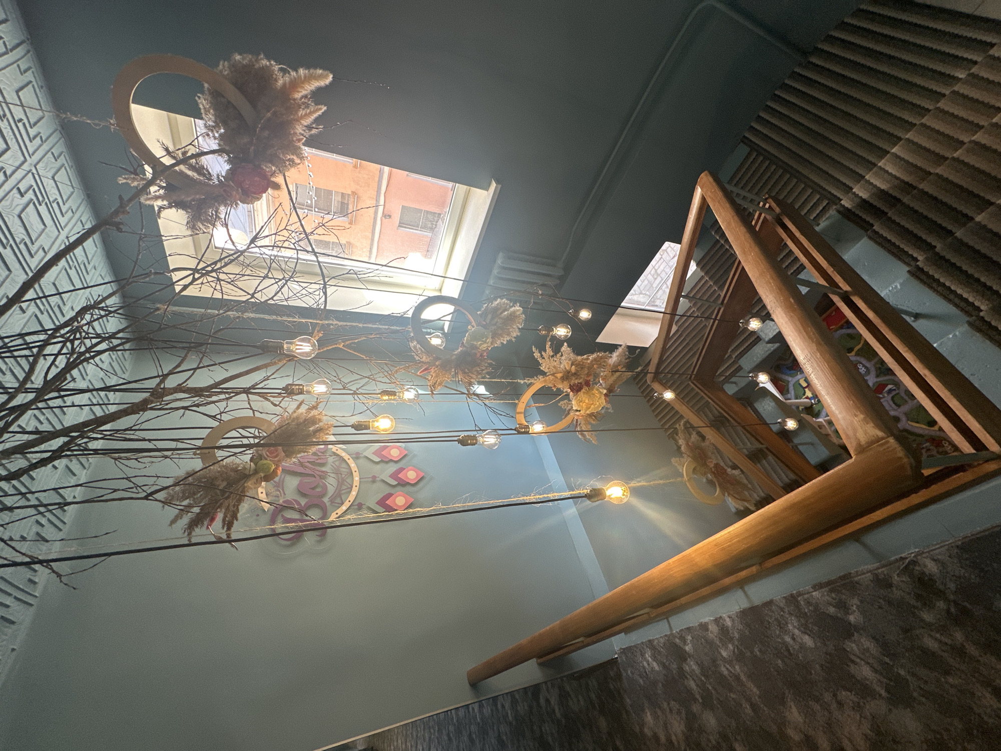 The image depicts an interior scene. The walls are painted in a muted teal color with decorative elements including a large circular emblem with floral motifs and geometric patterns. A wooden structure, possibly a partial staircase, is visible in the foreground. Suspended from the ceiling are branches adorned with decorative hoops and flowers. Light bulbs are strung between the branches, creating a whimsical effect. Through the windows, the facade of a building is partially visible. Given the decorative style and building details, the scene likely originates from an interior space in Saint Petersburg, Russia.