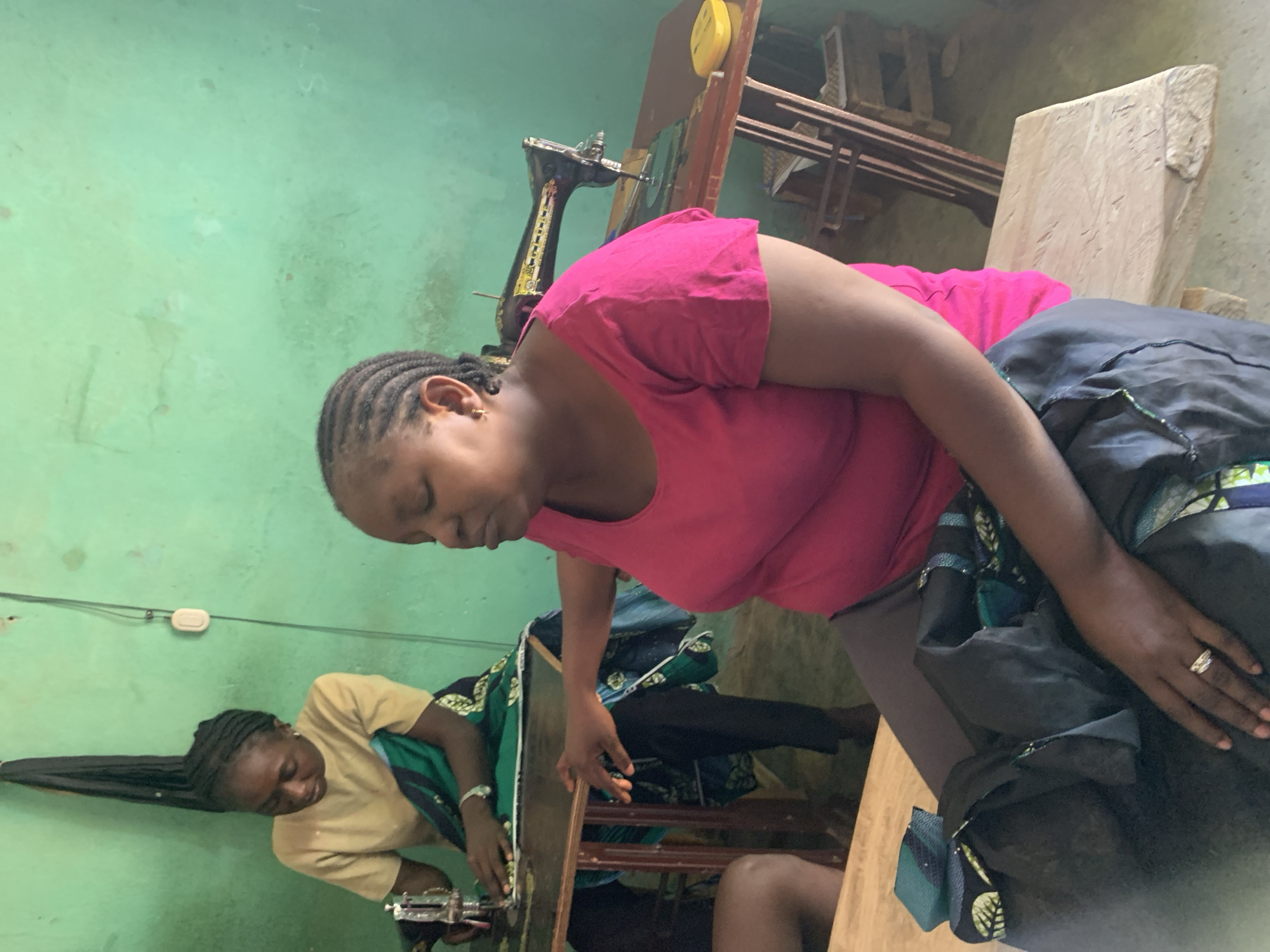 In Maigizo, Nigeria, two women are engaged in sewing. One woman, wearing a pink top, sits at a table with a sewing machine. She is looking down and appears to be working on a piece of fabric. The second woman, wearing a beige top, is also at a sewing machine. The room has green walls and appears to be a workshop, with a wooden table, and other sewing-related items.