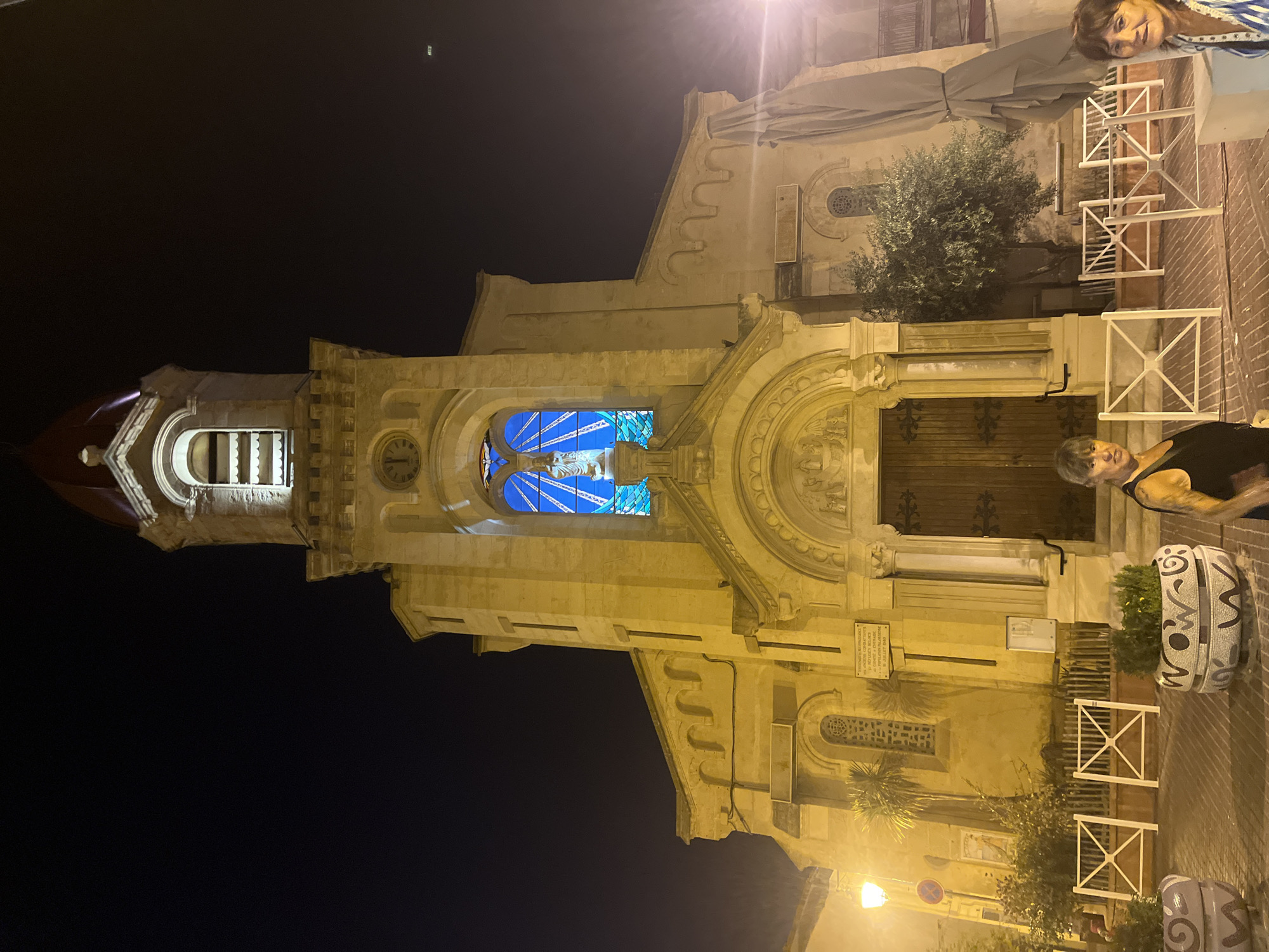 An exterior shot of the Église Saint-Pierre church in Palavas-les-Flots, France, at night. The church facade is illuminated with a warm yellow light, highlighting its architectural details, including arched doorways, windows, and a bell tower topped with a red dome. A stained-glass window depicting a figure, possibly Jesus on the cross, is visible above the main entrance, glowing with a vibrant blue light.
In the foreground, two women are partially visible. The woman on the left, facing the camera, has short dark hair and is wearing a black tank top. The woman on the right, also facing the camera, has shoulder-length brown hair and is wearing a striped top. They are standing in front of white decorative barriers and potted plants. The ground is paved with brick. Streetlights cast a warm glow on the scene. The background is dark, indicating nighttime.