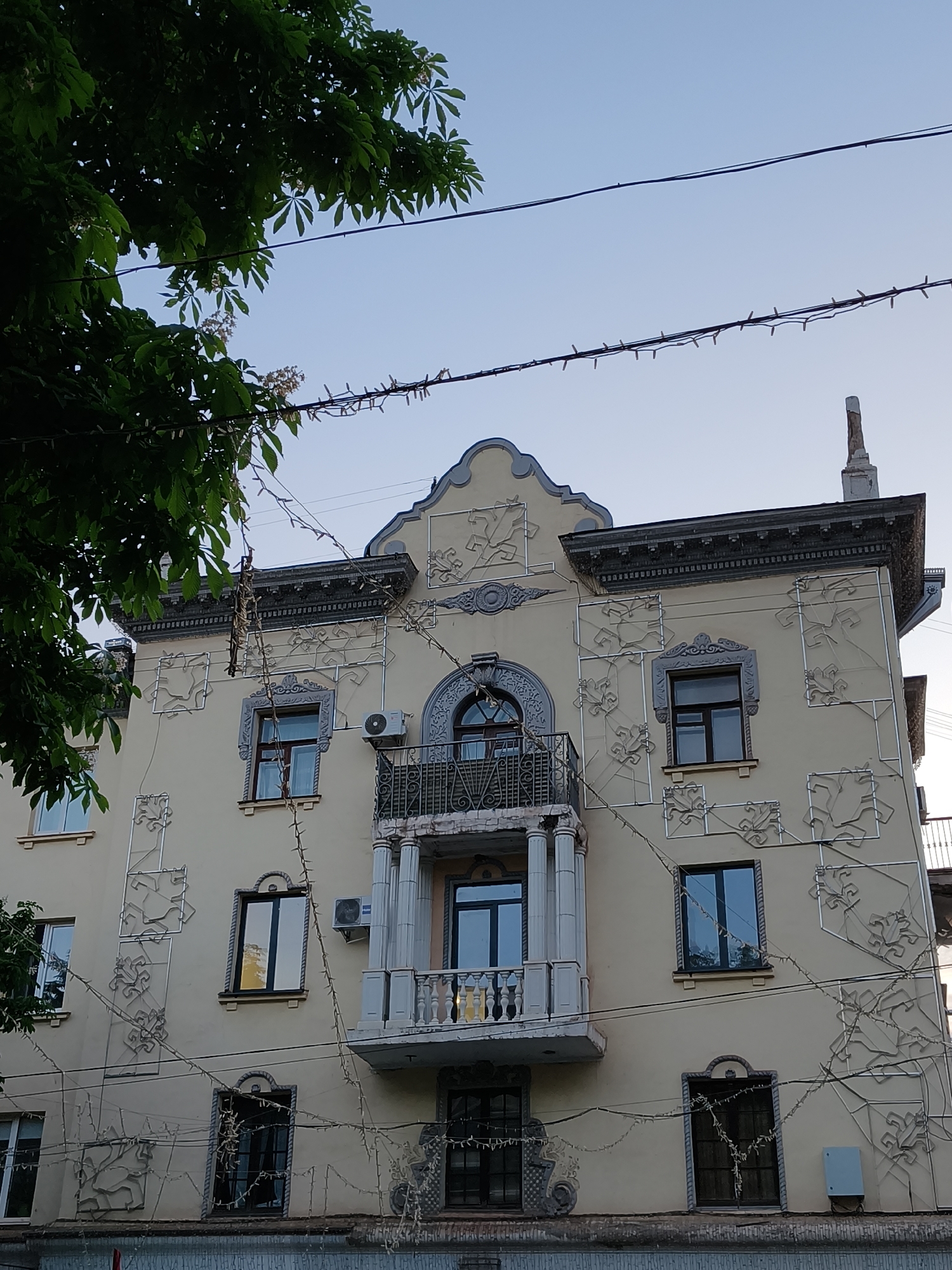 The image depicts a light yellow, ornate building in Kryvyi Rih, Ukraine. It is a residential building with a balcony on the second floor, bordered by two Corinthian-style columns. Above the balcony window is an arched window with ornate wrought iron detail. The building has several windows with dark frames, some reflecting the sky. Decorative plasterwork in geometric floral patterns adorns the façade. There are strings of lights hanging on the exterior of the building. Green tree foliage is visible on the left side of the image, and electrical wires run across the top against a light blue sky.