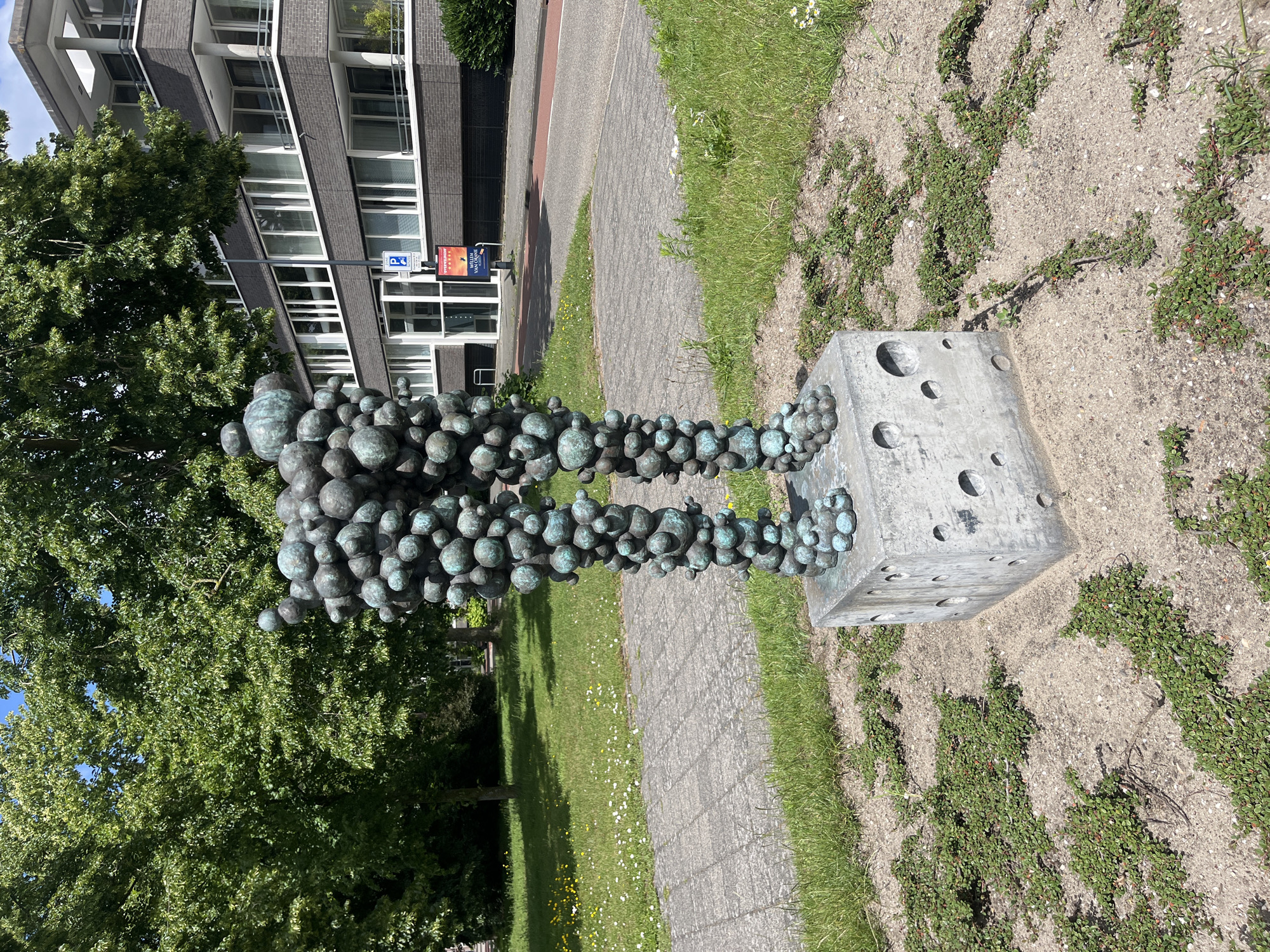 A bronze sculpture of two legs stands on a concrete cube in a grassy area. The legs are made of many spheres of varying sizes. The concrete cube has circular holes in its sides. In the background, there is a sidewalk, trees, and a modern building with many windows. The sky is blue and overcast.