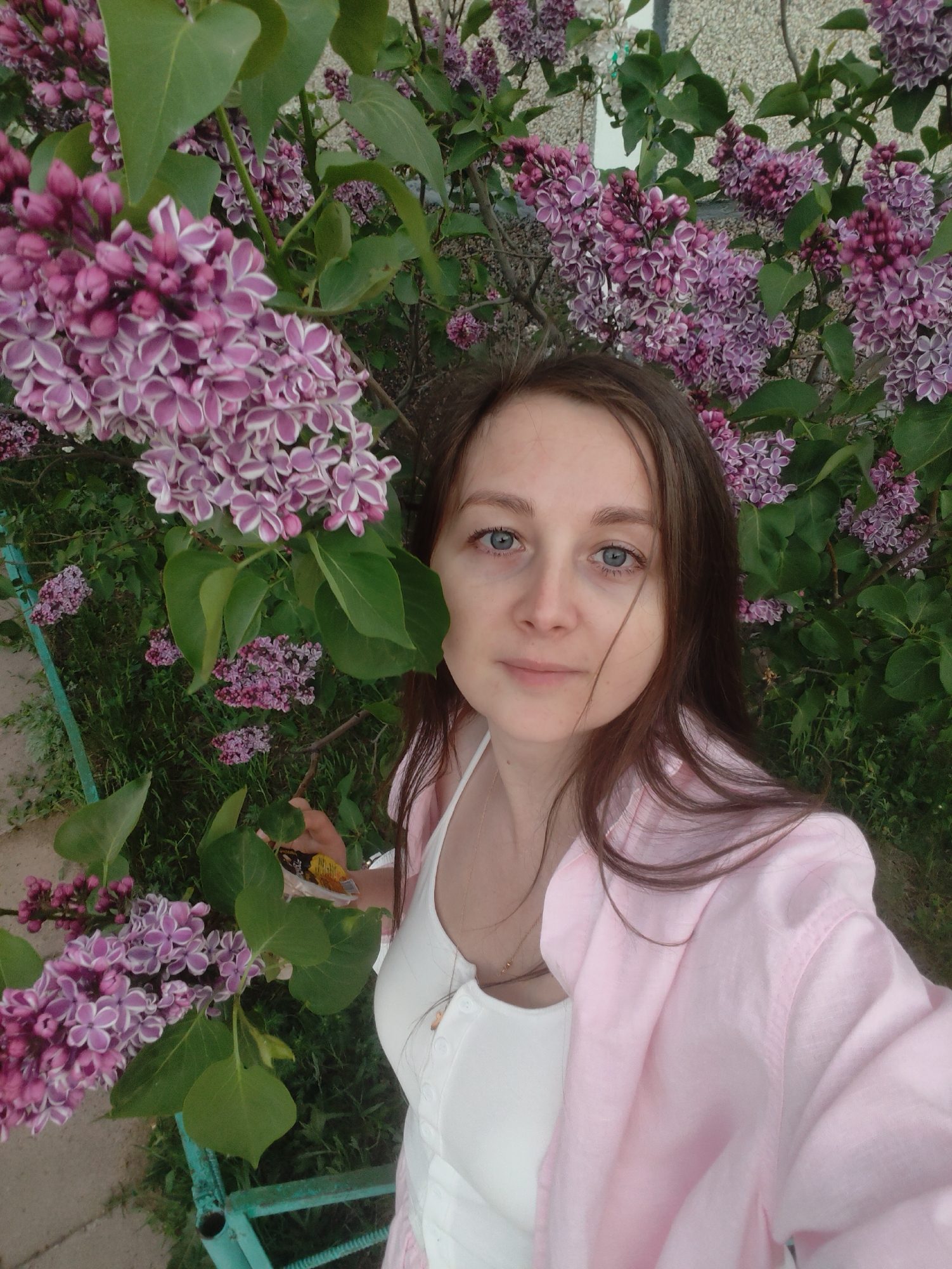 The image shows a woman with brown hair and blue eyes taking a selfie next to a lilac bush. She is wearing a white top and a light pink shirt. The lilac bush is full of purple and white flowers and green leaves. The background is blurry, but it appears to be a building. The location is Unknown, Unknown.