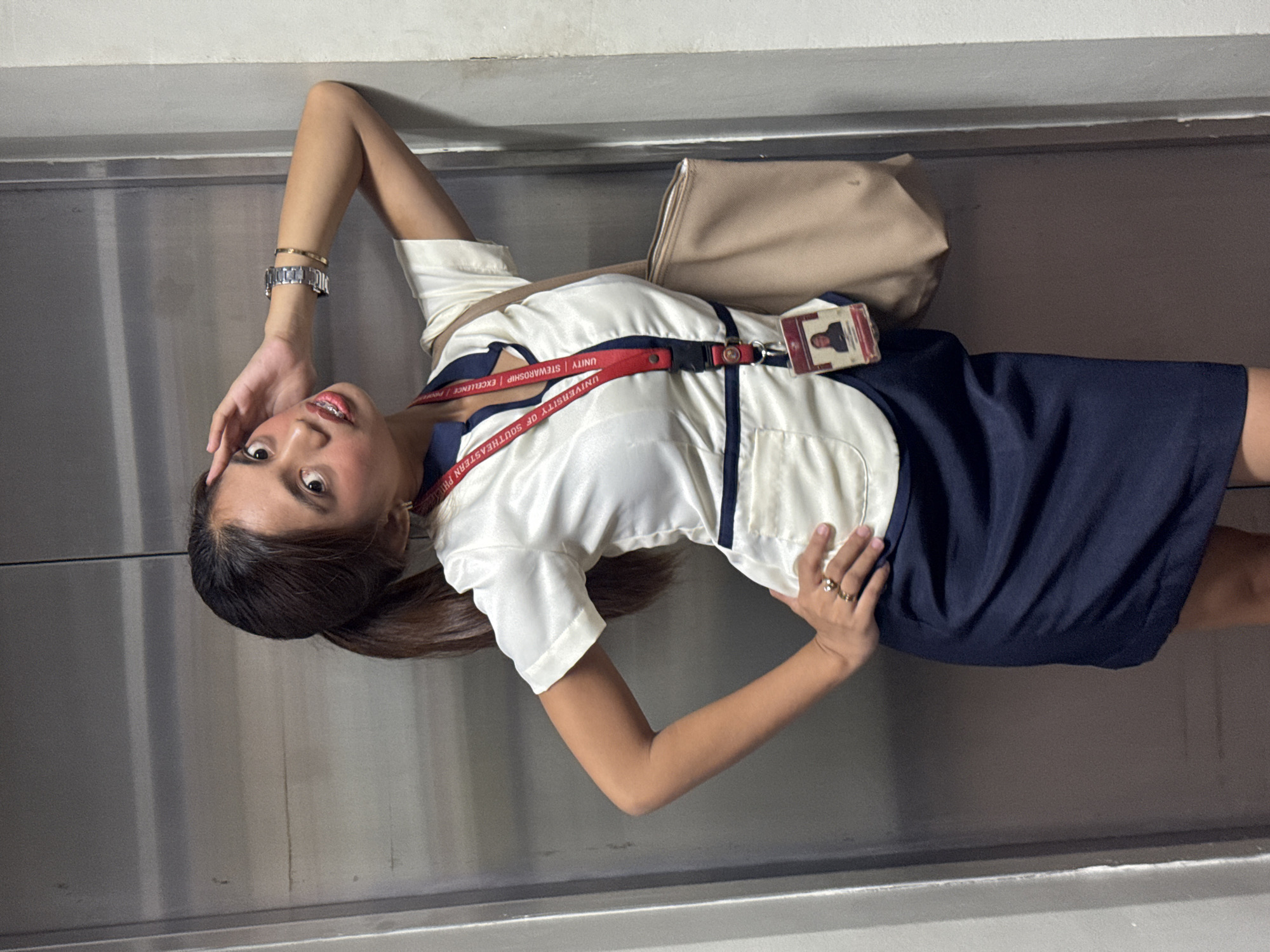 A young woman with dark hair pulled into a ponytail stands in front of a metallic elevator door. She is wearing a white collared shirt with navy trim and a navy skirt. A lanyard with an ID card hangs around her neck. She has a light-colored tote bag slung over her shoulder. Her left hand is raised to her forehead, and her right hand is on her hip. She is wearing jewelry on her wrists and fingers. The background appears to be an indoor hallway with a textured wall to the right of the elevator. There are no discernible landmarks indicating a specific location within Davao City, Philippines.
