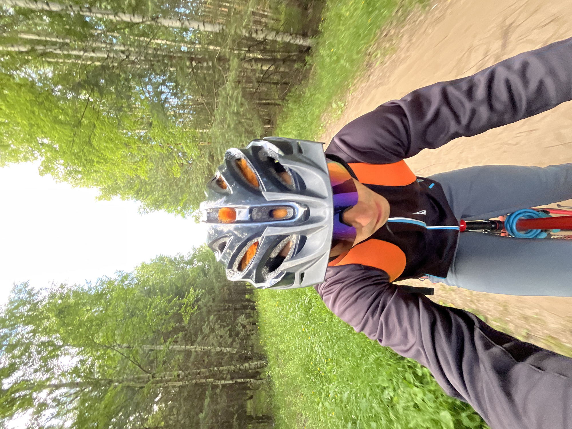 The image shows a first-person perspective of a person cycling on a dirt path through a forest. The subject is wearing a silver and black bicycle helmet, sunglasses with purple and orange lenses, a dark grey long-sleeved cycling jersey with orange accents, and grey cycling shorts. Their arms are visible, one of which is holding onto bicycle handlebars, and a blue cable lock is attached to the bike.
The scene is a forest with lush green trees and grass alongside the path. The path itself is a light brown dirt track. The lighting suggests it is daytime, with bright sunlight filtering through the trees, creating some dappled light and shadow. The overall atmosphere appears to be peaceful and active, indicative of recreational outdoor activity.
There is no visible text in the image. The location context provided is Chaikovsky, Russia.
