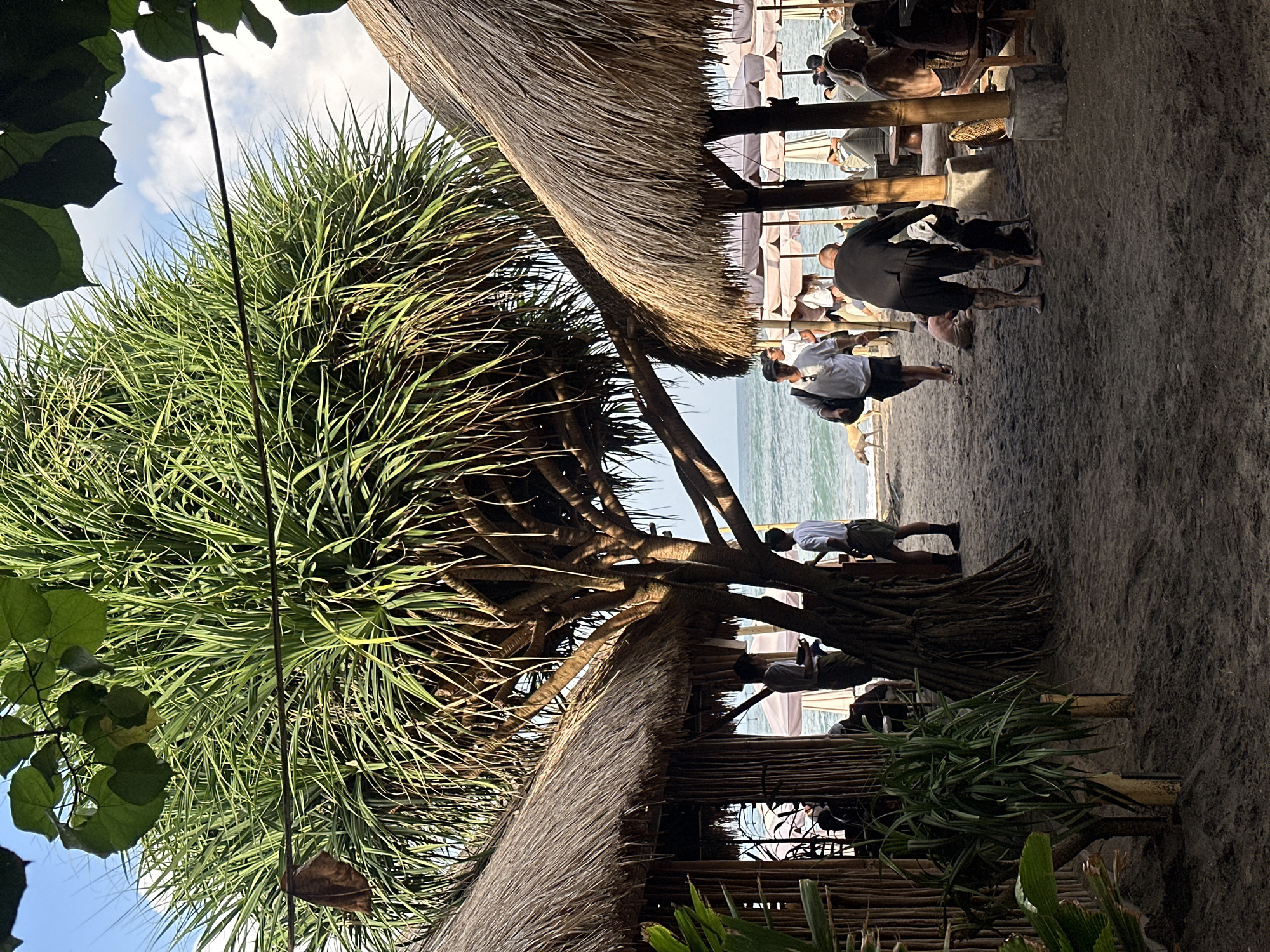 The image depicts a sandy beach scene in North Kuta, Indonesia, characterized by a large pandanus tree providing shade. Two thatched-roof huts are visible, one on the left and one on the right, with bamboo structures. People are scattered across the sand, some walking and others seated in chairs or on lounge-like seating under umbrellas. A man in a black shirt and shorts is interacting with a black dog. Another person, partially visible, is lying on the sand. In the background, the ocean is calm with a few waves, and the sky is partly cloudy. Several other individuals are walking along the beach, and a light-colored dog is also present. The overall atmosphere suggests a relaxed beachside setting.