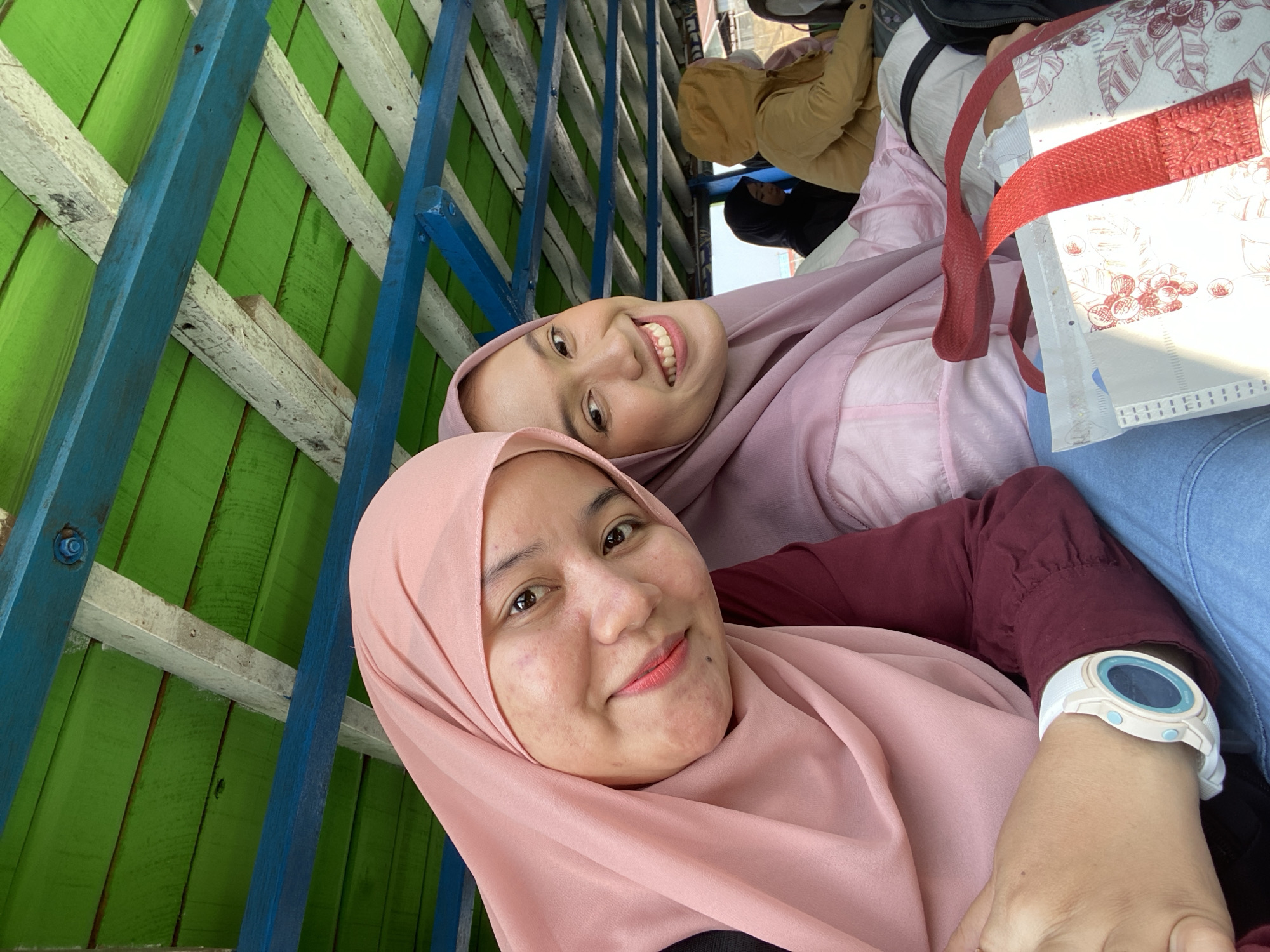 Two women are taking a selfie on a boat with a green and blue painted wooden roof. The woman on the left is wearing a pink hijab and a maroon top, and a white watch on her left wrist. The woman on the right is wearing a light purple hijab and a light pink top. The boat is likely operating on the waters of Banjarmasin, Indonesia, known for its riverine transportation. In the background, other passengers are visible, including one wearing a yellow hooded jacket and another in a black hijab. A reusable shopping bag with a red handle and a patterned design is in the foreground.