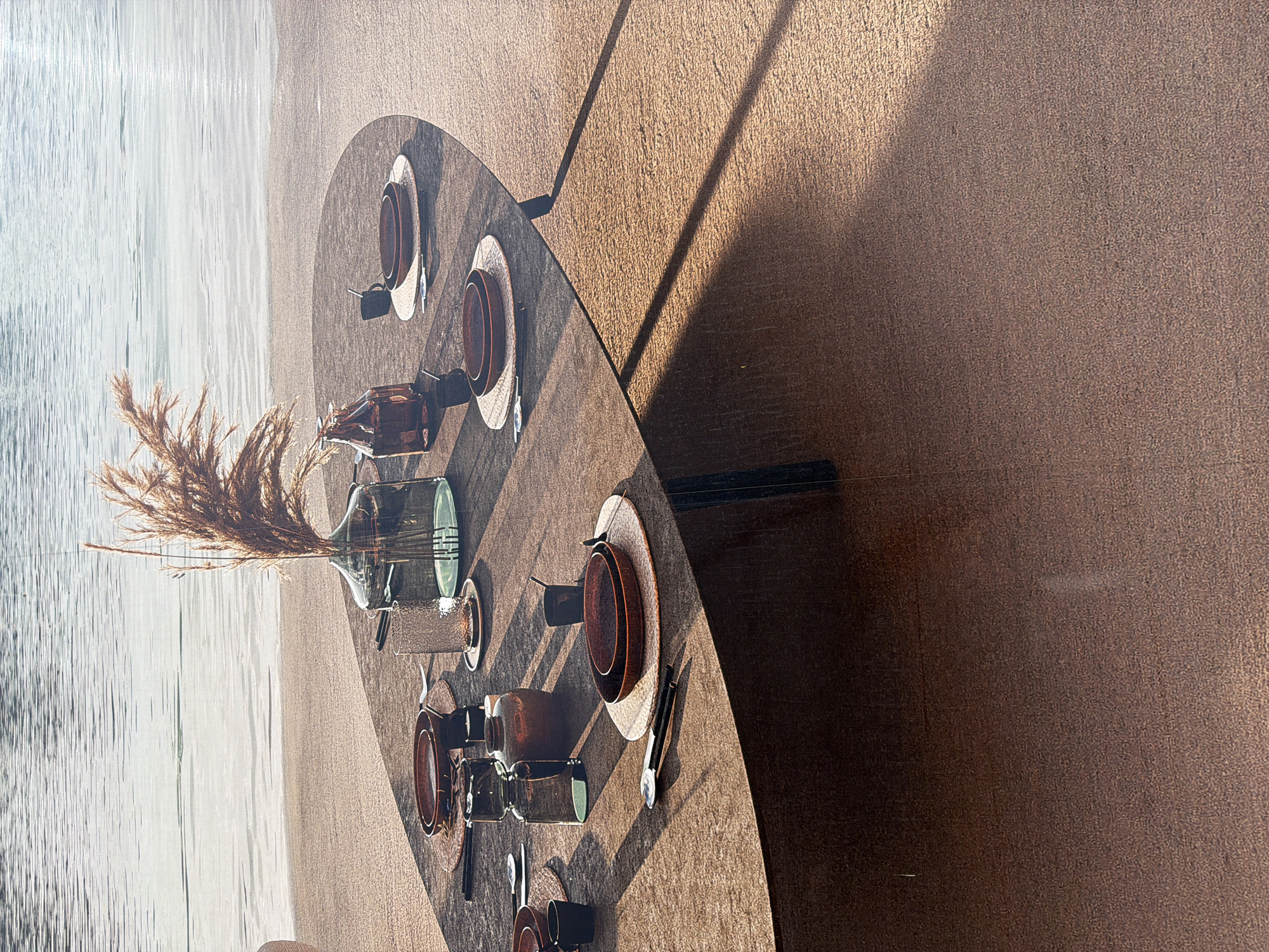 The image depicts an outdoor dining setting next to a body of water, likely a river or lake, given the gentle ripples and the sandy shoreline. The scene is situated in Mol, Belgium. A large, oval-shaped table, made of a textured, cork-like material, is set for a meal. The table has a warm, earthy tone.
On the table, there are several place settings. Each setting includes a round, brown placemat, topped with a larger, shallow brown bowl and a smaller, deeper brown bowl nested within it. Cutlery, consisting of a knife and fork, is placed on the left side of each setting. There are also small, dark mugs or cups at some settings.
A prominent feature of the table is a large, clear glass vase filled with dried, feathery grasses, adding a natural, rustic element to the arrangement. Beside the vase, there are two dark brown glass bottles, one taller than the other. Another smaller, opaque brown vase or jar is also present on the table. A textured, clear glass tumbler sits on a coaster.
The table is positioned on a surface that appears to be made of the same cork-like material as the tabletop. Sunlight casts strong shadows across the table and the surrounding surface, indicating a bright, sunny day. The background shows the expanse of the water and the sandy bank. No people are visible in the image.