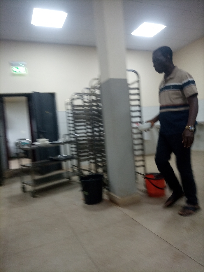 A man stands in a commercial kitchen. He wears a striped shirt, dark pants, and sandals, holding a white bowl. Stainless steel baking racks line a wall next to a gray pillar. A rolling cart with supplies and trays sits near an open doorway. Two trash cans, one black and one orange, are positioned on the tiled floor. Overhead, square fluorescent lights illuminate the room. A green exit sign glows above the doorway. The image is somewhat blurry, indicating movement.