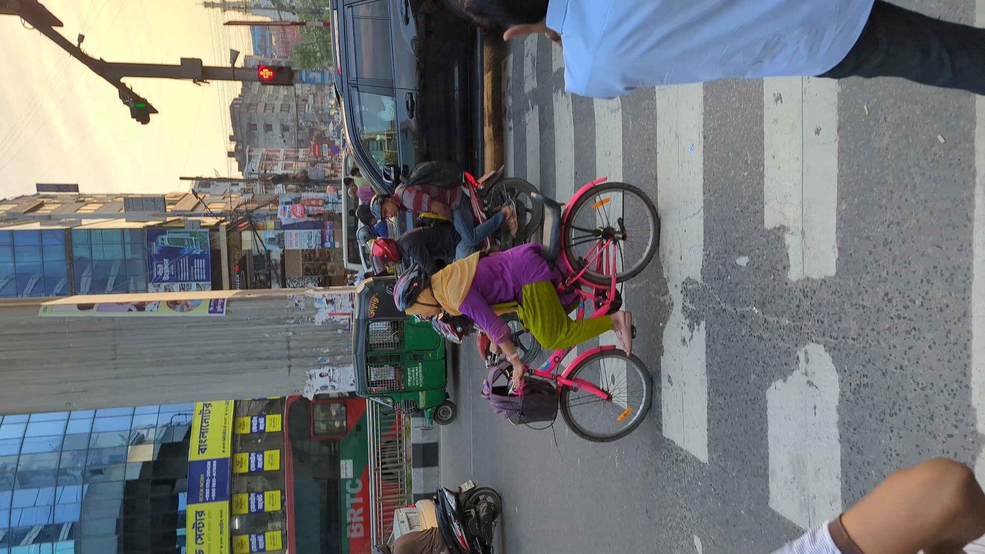 The image depicts a bustling street scene in Dhaka, Bangladesh. A person in a purple top and green pants is riding a pink bicycle across a zebra crossing. Several other people are also crossing the street, some on bicycles and motorcycles. A black van is parked nearby, and a green auto rickshaw is visible in the background. A red bus with the BRTC logo is partially visible on the left side of the image. The street is lined with buildings displaying various advertisements and signage in Bengali. A traffic signal shows a red light.