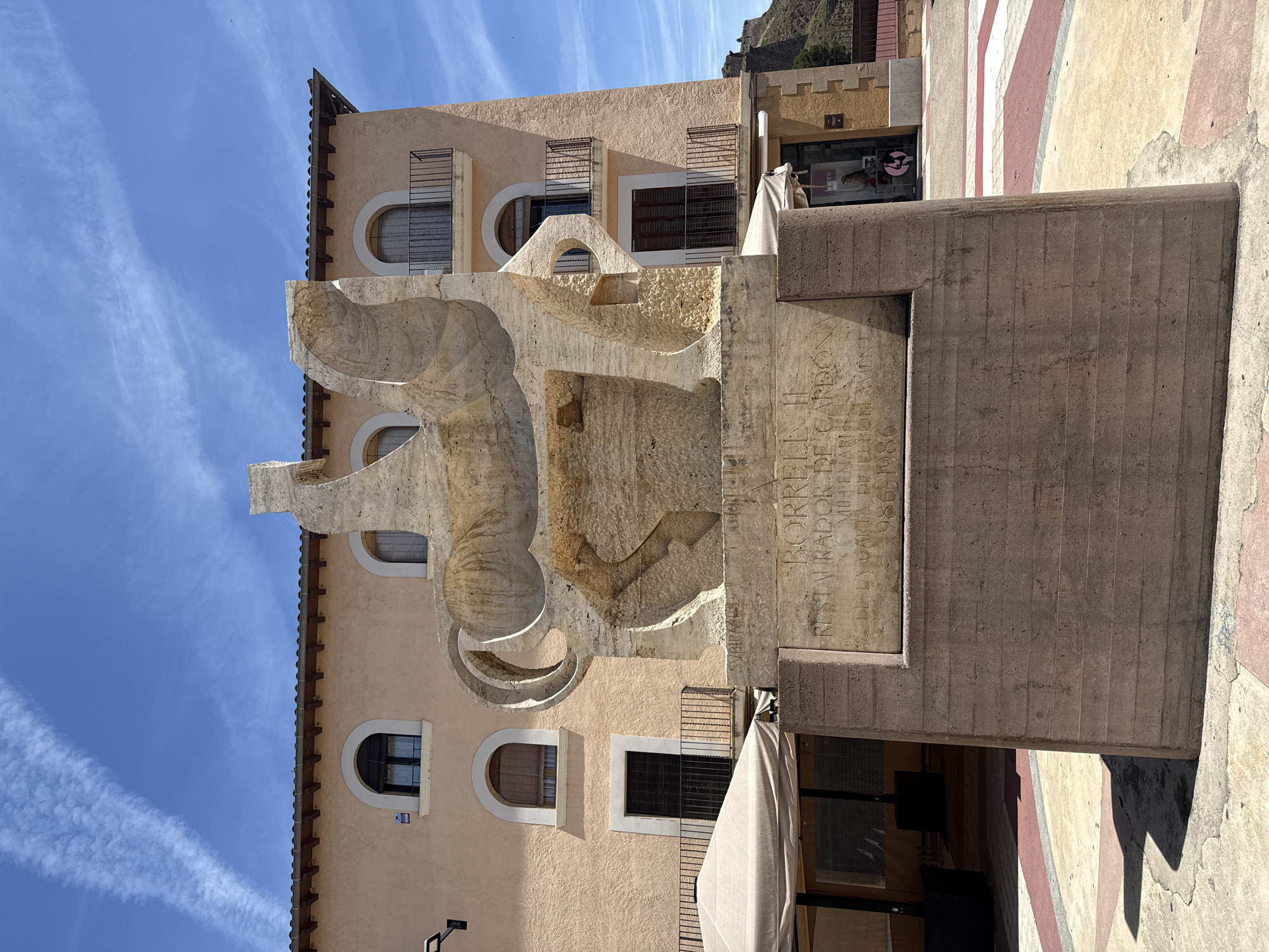 An outdoor, eye-level, medium shot captures a stone sculpture of a horse and rider in the city of Cardona, Spain. The sculpture is light beige and features abstract, geometric shapes. The horse's body is a solid mass with carved-out sections, while the rider is depicted as a simplified, upright figure. The sculpture rests on a larger, reddish-brown stone pedestal with inscribed text and a carved date, "1986". Behind the sculpture, a pale orange, multi-story building with arched windows and black balconies stands under a bright blue sky with wispy white clouds. To the left of the sculpture, a light beige awning covers an outdoor seating area. The ground in front of the sculpture is paved with colored concrete sections. The scene is bathed in bright sunlight, casting distinct shadows on the ground.