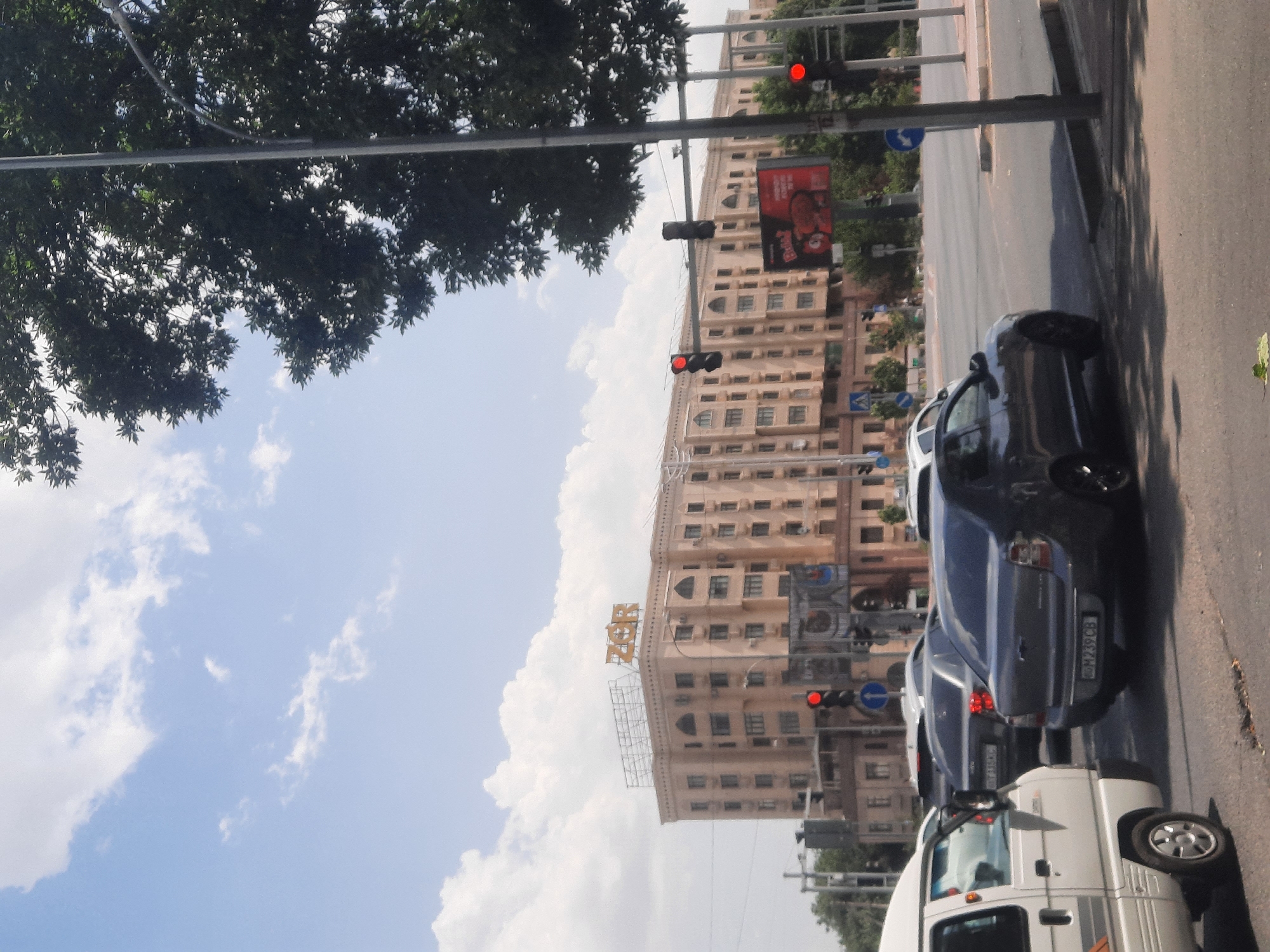 In Tashkent, Uzbekistan, a street scene unfolds under a partly cloudy sky. The focal point is a large, multi-story building with a tan facade and numerous windows. Atop the building, a sign reads "ZOR" in large, yellow, three-dimensional letters. Several cars are visible, including a white van in the foreground on the left, a dark gray sedan with license plate "M239CB" in the middle of the road, and other vehicles partially obscured. Traffic lights show red, indicating a stop. Trees with dense green foliage frame the right side of the image, and a lamppost stands tall. A billboard with a red background and an image of a pig is also present. The overall impression is of a busy intersection in a city.