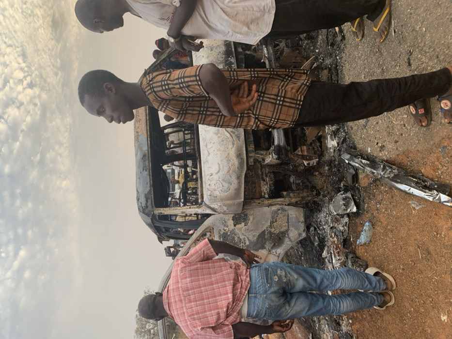 A burnt-out vehicle, possibly a bus, is surrounded by onlookers. Three men are in the foreground: one in a white shirt, another in a brown and black checkered shirt, and the third in a red plaid shirt and jeans. The vehicle's exterior is heavily charred, with visible interior details and potential passengers. Debris litters the ground around the vehicle. The sky is overcast, suggesting a somber or serious atmosphere.