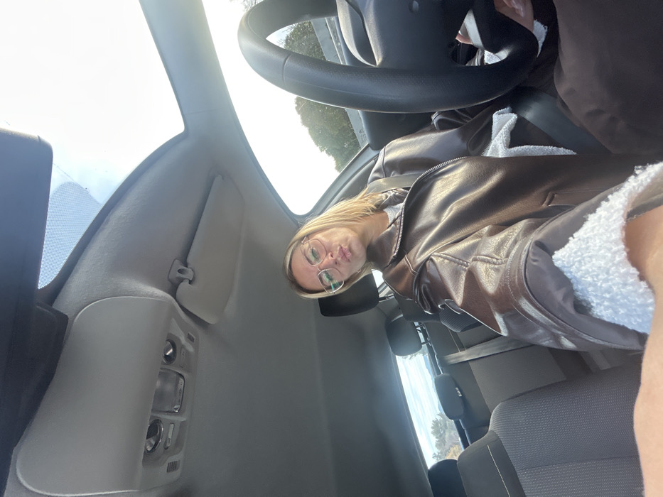 A female individual with medium-length blonde hair and clear-framed eyeglasses is seated in the driver's position of a passenger vehicle, taking a selfie. She is wearing a brown, leather-look jacket with visible white sherpa lining at the cuffs and collar, over a white inner layer. Her seatbelt is fastened across her chest, and her lips are pursed. The vehicle's interior features a light grey headliner, an overhead console with interior lights and a sun visor. A panoramic sunroof is visible above. A black steering wheel is partially visible on the right side of the frame. The background through the windows shows a bright outdoor setting with trees and distant vegetation, indicative of a road or highway environment. The scene is situated in Villeneuve-Loubet, France.