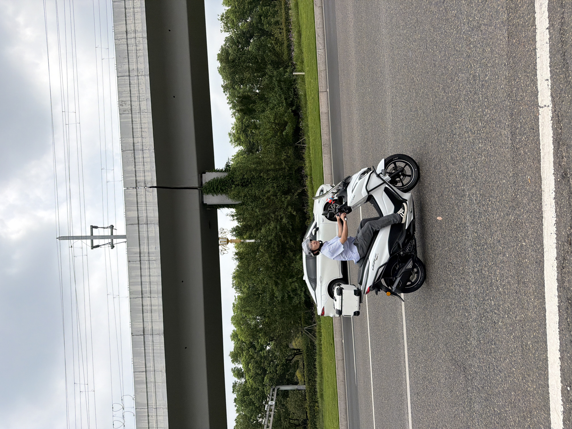 A man rides a white scooter on an asphalt road in Jiangjin, China. The man wears a striped shirt, grey pants, a helmet, and sunglasses. The scooter has a white storage box on the back. In the background, a white car is parked near a line of trees. An elevated highway or overpass with power lines runs overhead. The sky is cloudy.