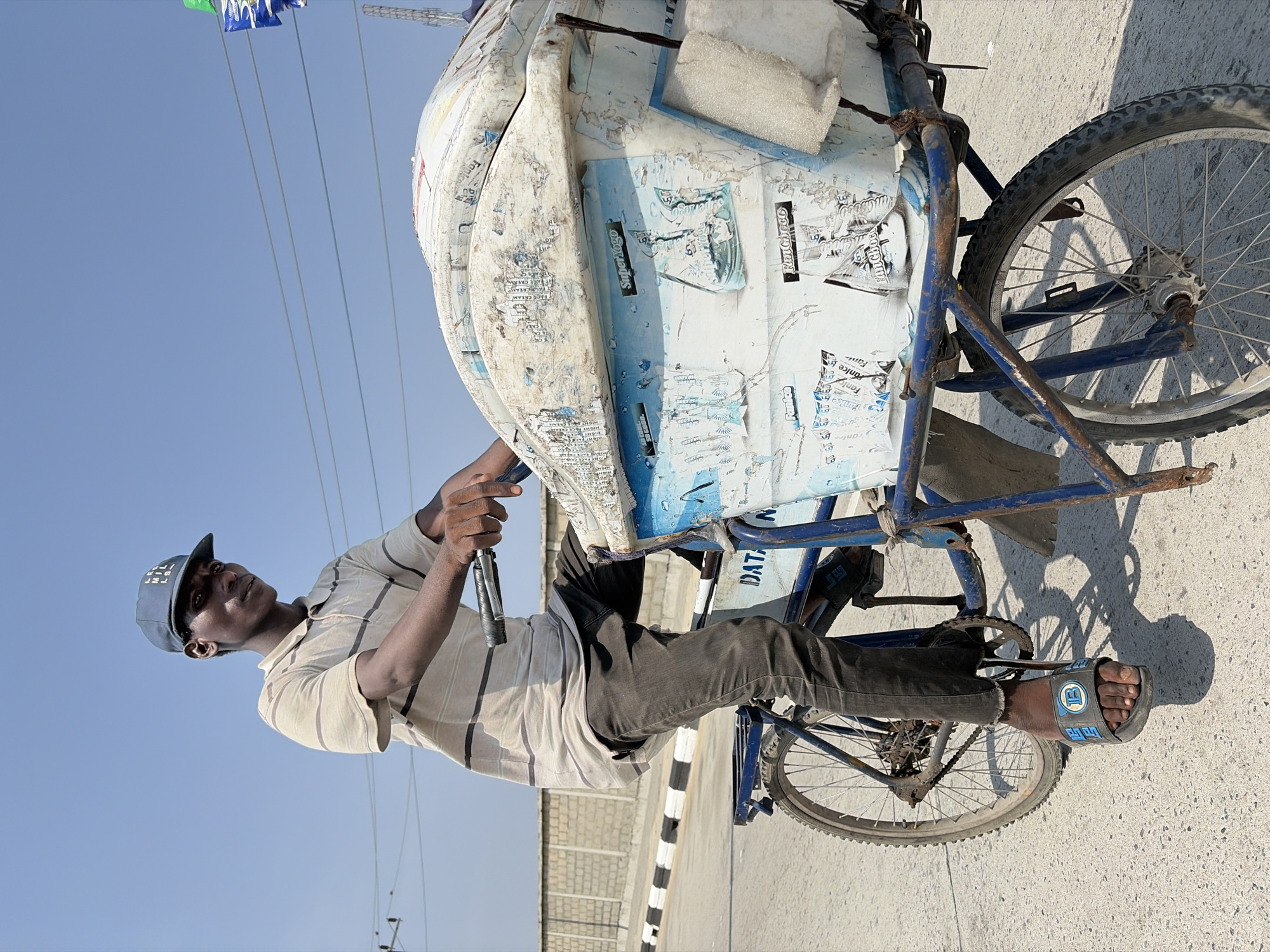 A man in Igboefon, Nigeria, is riding a three-wheeled bicycle. The vehicle has a large, box-like cooler or cart attached to the front, covered in stickers and advertisements. The man is wearing a blue baseball cap, a striped beige shirt, and dark pants. He is pedaling the bike with his right foot in a sandal. The setting appears to be a street in Igboefon, with a clear blue sky overhead and a partially visible wall and power lines in the background. The bicycle shows signs of wear and rust.