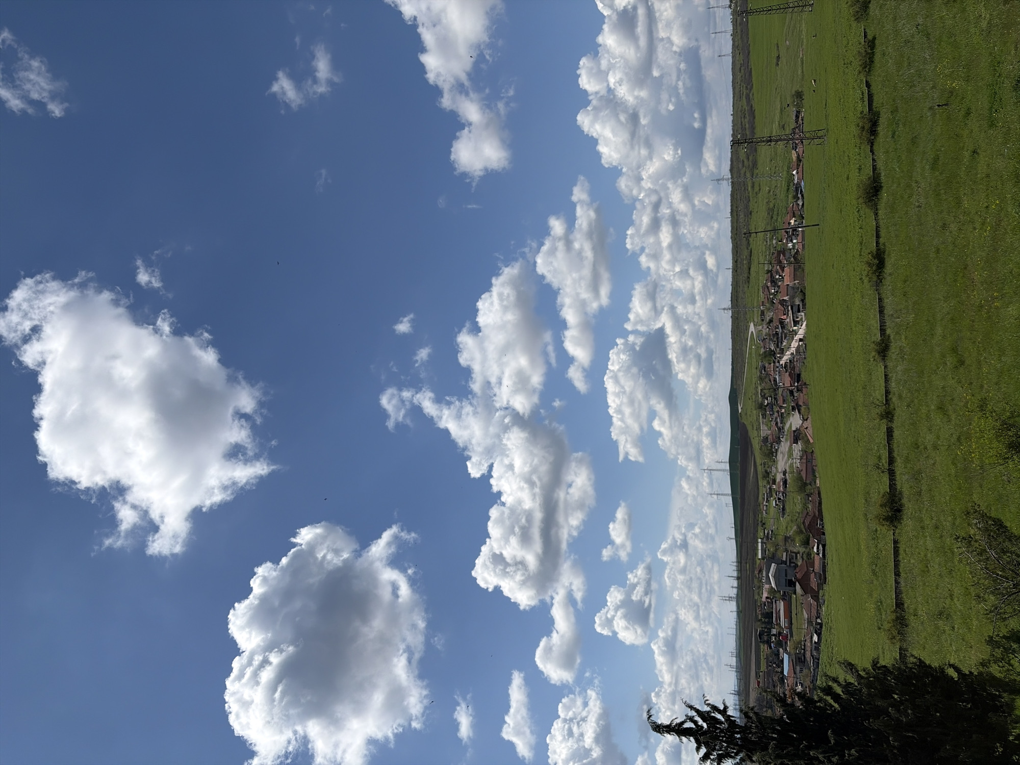 The image depicts a rural landscape under a blue sky dotted with fluffy white clouds. A field of green grass stretches across the right side of the image. A small village or town is nestled between the field and the sky, marked by the reddish-brown roofs of the buildings and the presence of power lines. The location is unknown.