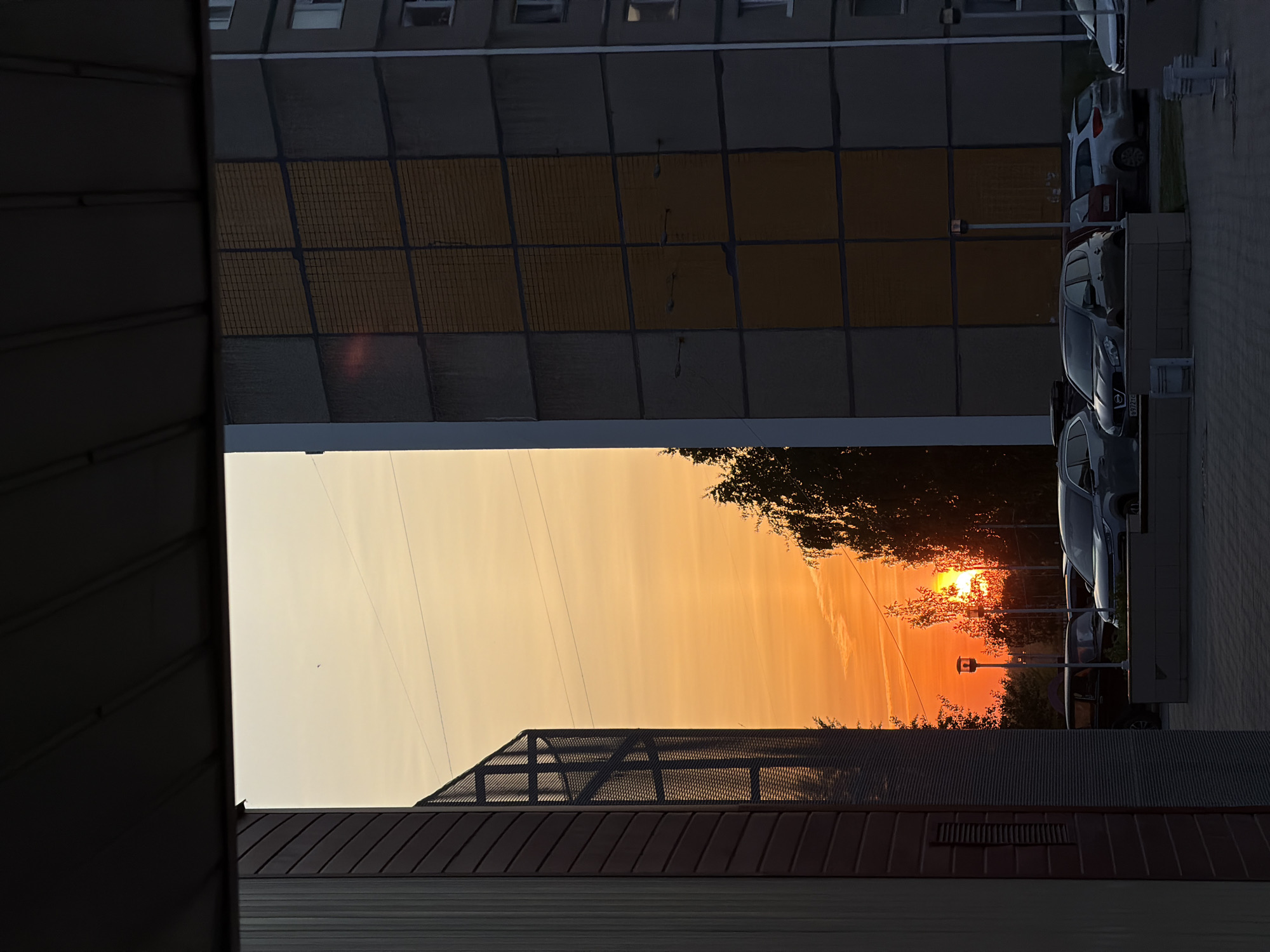 The image, taken in Chaikovsky, Russia, shows a sunset framed by buildings. On the left is a dark building with vertical siding, and below, a structure with a metal grate. Above, is a tall gray building with a section of yellow grid panels. To the right, cars are parked along a curb. The sunset casts an orange glow in the sky visible between the buildings. Trees silhouette against the bright sun. There are powerlines crossing the sky.