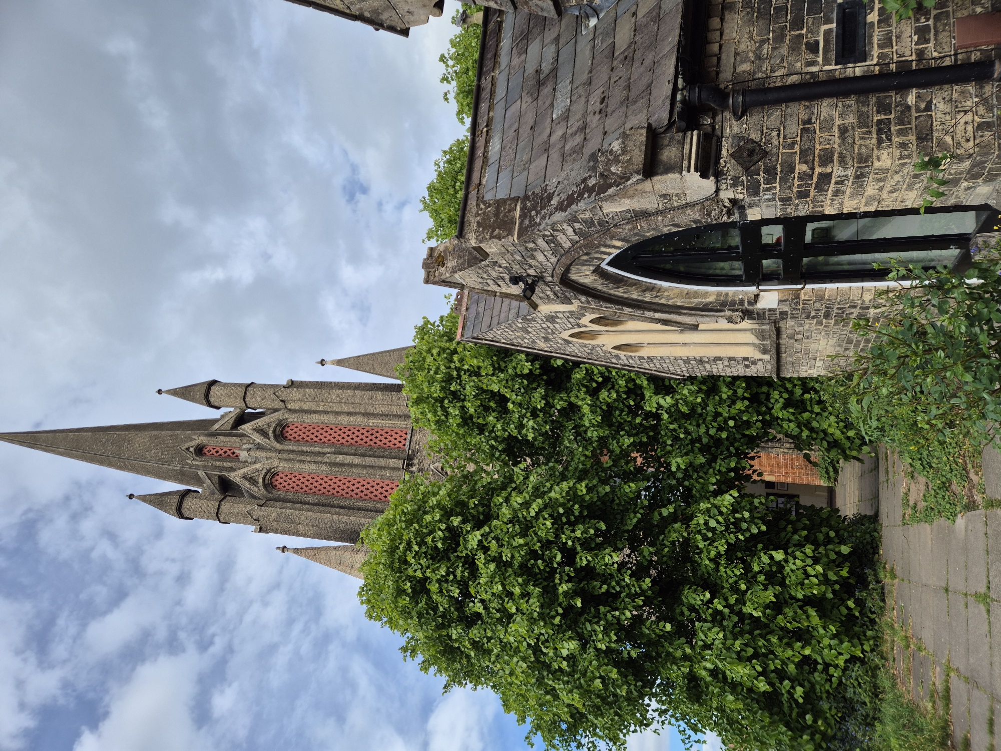 An exterior shot depicts a Gothic church tower, adjacent to a stone building with a rounded window and slate roof, partially obscured by lush green foliage, under a cloudy sky. A paved walkway extends into the scene, suggesting an urban environment. The location is West Suffolk, United Kingdom.