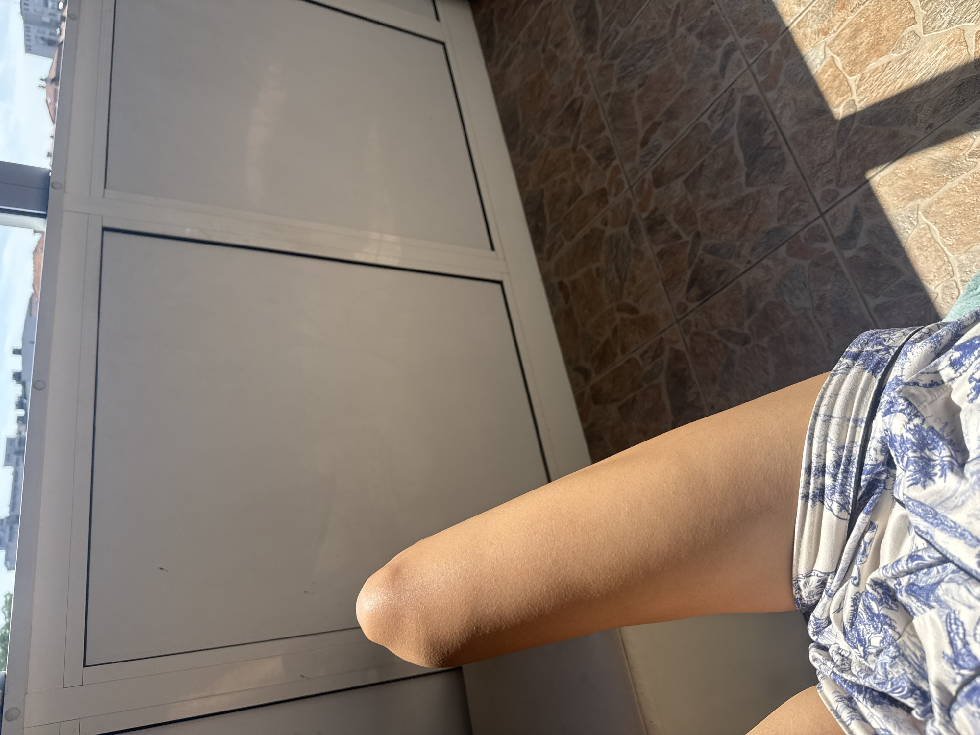 The image shows a person's legs in shorts on a tiled floor, likely on a balcony or enclosed porch with a view of buildings. The light suggests it is daytime. The location is the city of Plovdiv, Bulgaria.