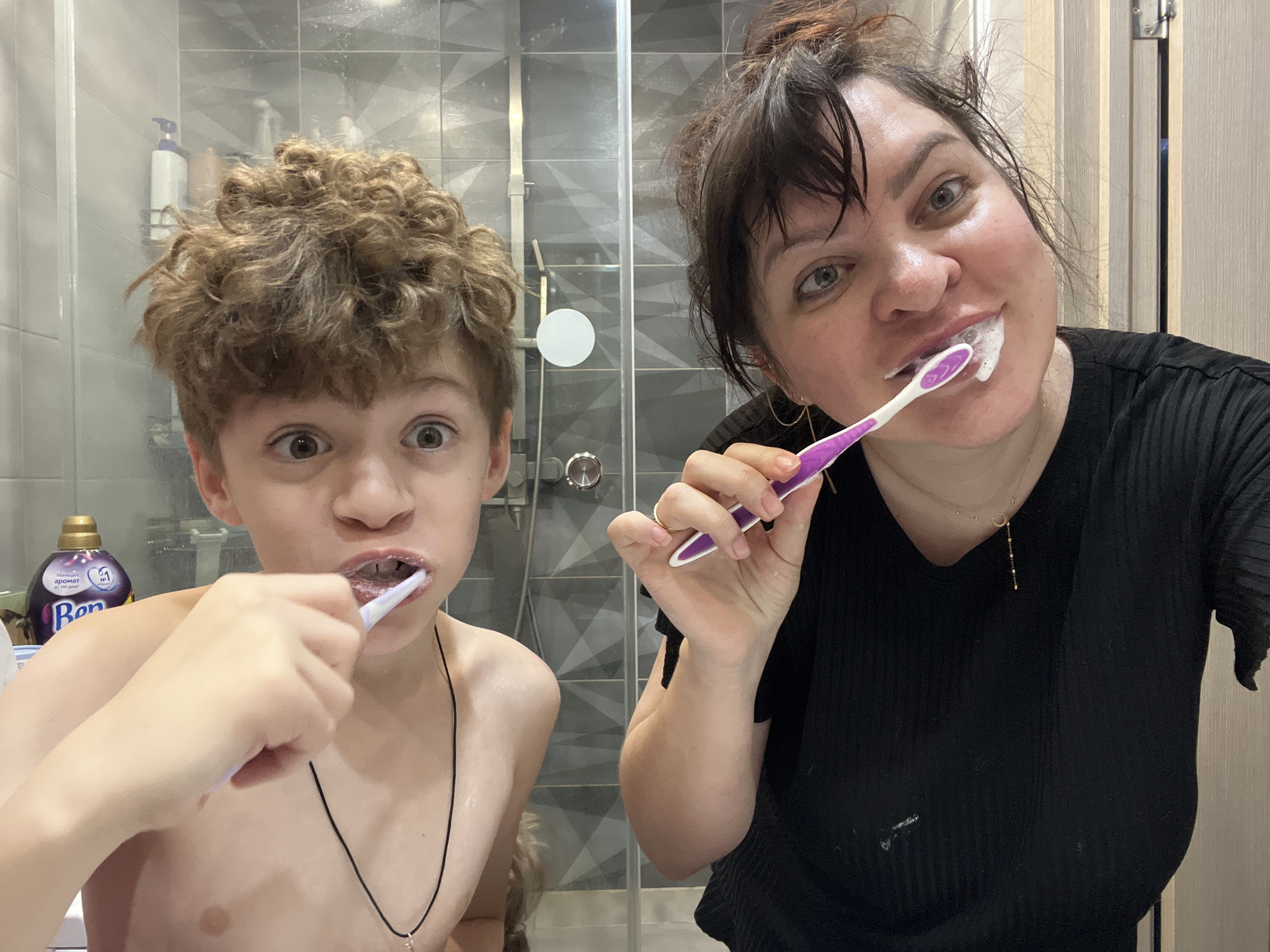 The image depicts two individuals, an adult female and a male child, engaged in oral hygiene within a bathroom located in Noviy, Russia.
The adult female, positioned on the right, has dark hair pulled back loosely, fair skin, and light-colored eyes. She is smiling directly at the camera with toothpaste foam visible around her mouth, while holding a purple and white toothbrush in her right hand. She wears a black ribbed V-neck top, a gold-colored necklace with a small pendant, and a ring on her left ring finger. A smudge of white substance, consistent with toothpaste, is present on her shirt.
The male child, positioned on the left, is shirtless with light brown curly hair and fair skin. He also looks directly at the camera with wide, surprised eyes. He holds a white toothbrush in his right hand, brushing his teeth, with dark residue visible within his mouth. He wears a thin black cord necklace with a small pendant.
The background shows a modern bathroom environment featuring grey geometric patterned tiles and a glass shower enclosure. Behind the shower glass, several bottles are visible, including a prominent purple bottle identifiable as "Vernel" fabric softener with Russian text ("Vernel аромат №1"). A light-colored wooden door frame is partially visible on the far right. The scene is illuminated by artificial indoor lighting.