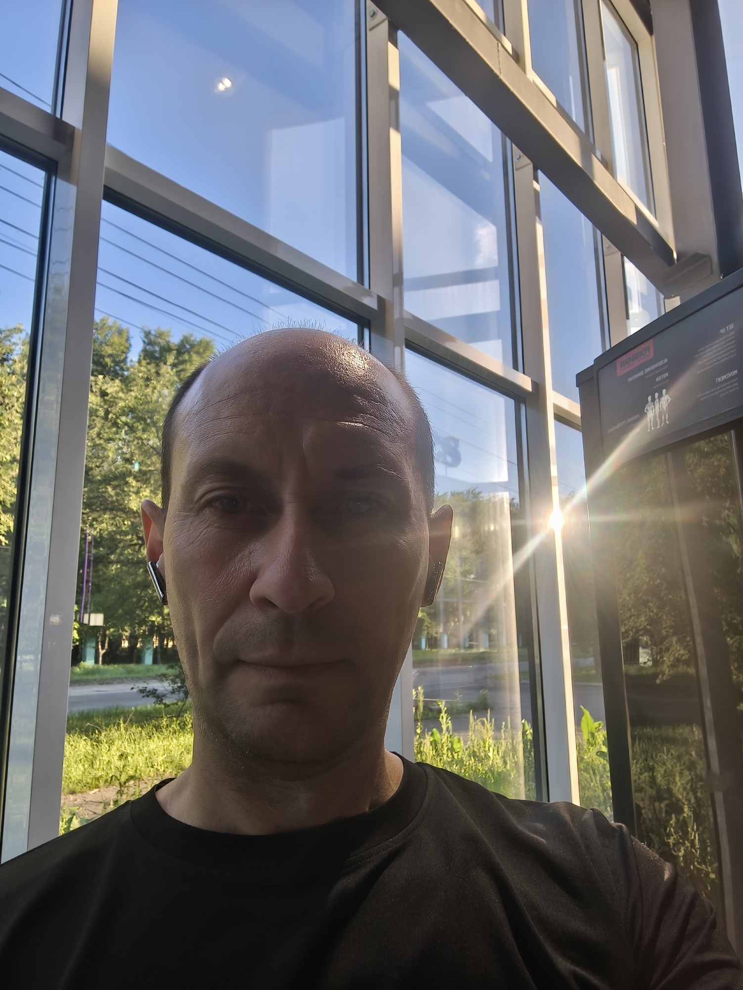 A bald man, wearing a black t-shirt and an earbud, is taking a selfie indoors, near large windows. The windows reflect the blue sky and trees outside, with the sun creating a lens flare. A digital display, possibly for exercise equipment, is visible to the right of the man. The man's face is lit from the side by the sun. The scene is likely in Komsomolsk-on-Amur, Russia, given the prompt.