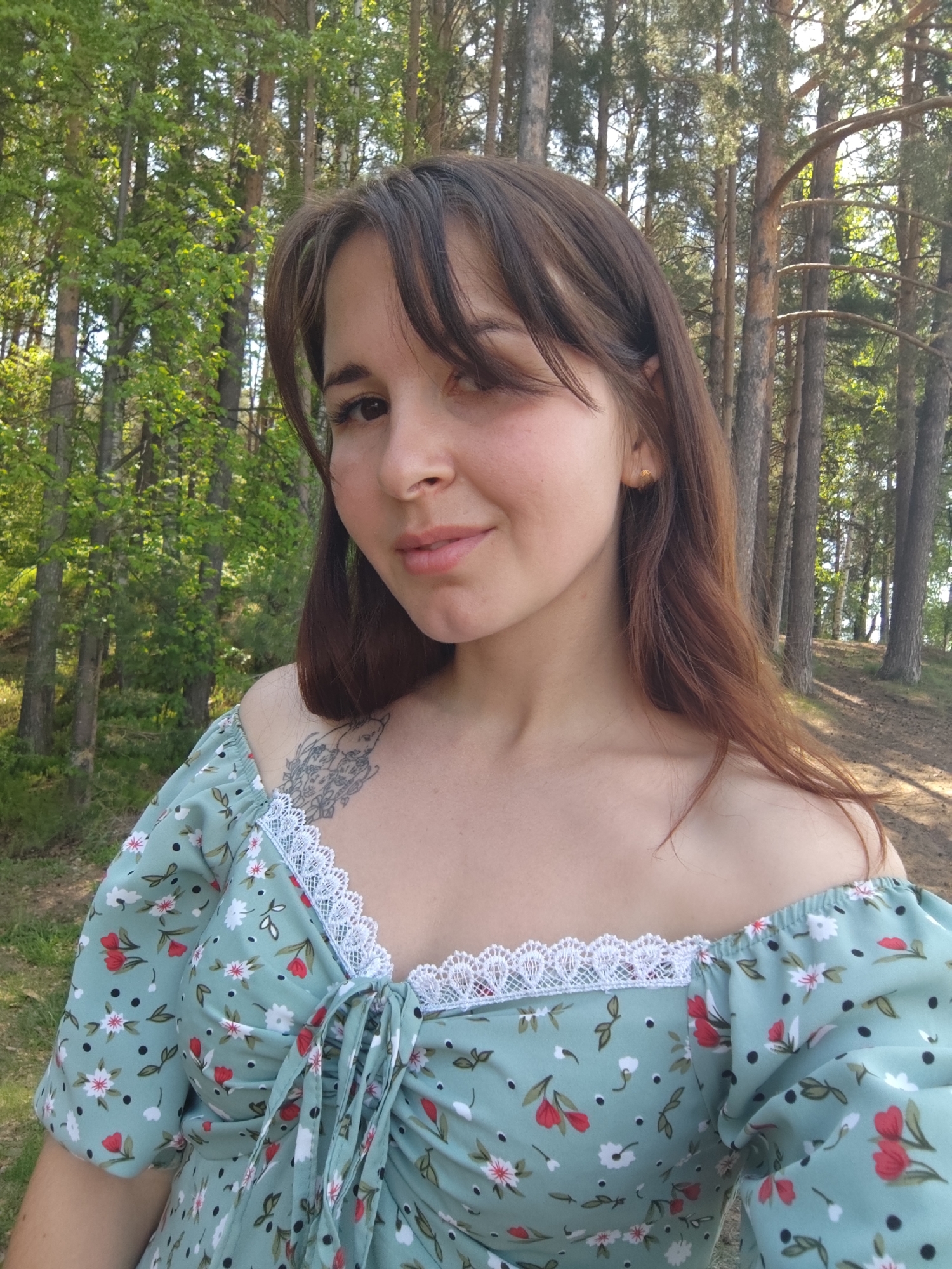 A young woman stands in a forest in the city of Chaikovsky, Russia. She has brown hair with bangs and wears a floral dress with white lace trim. A tattoo is visible on her left shoulder. The background features tall trees with green foliage. The woman is facing the camera with a slight smile.