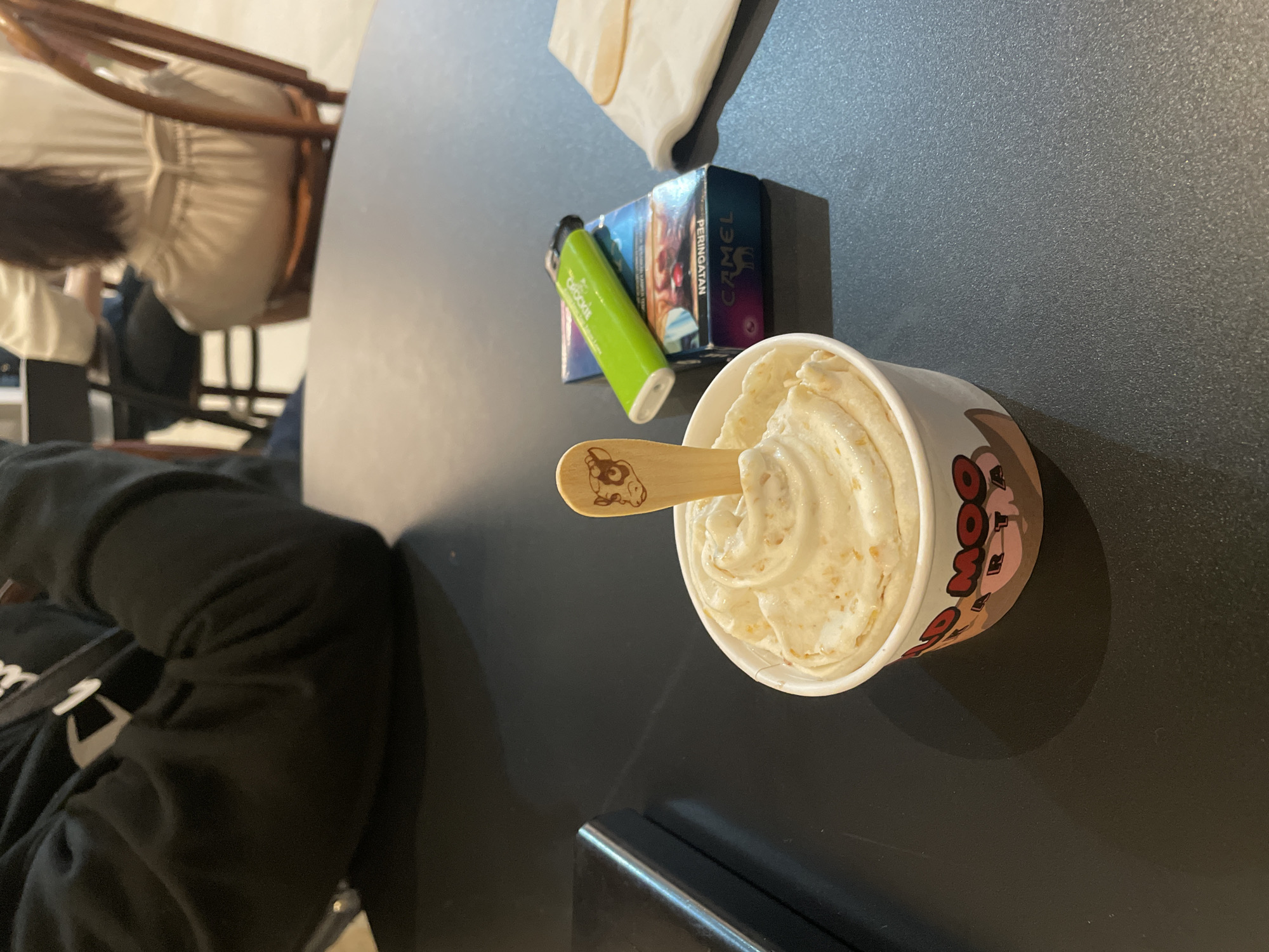 A high-angle shot depicts a cup of ice cream with a wooden spoon on a dark-colored table. Beside it are a box of Camel cigarettes and a green vape pen. In the background, a partial view shows a person seated on a chair. The location appears to be in North Jakarta, Indonesia, indicated by the "PERINGATAN" text on the cigarette box, which is a warning label used in Indonesia.