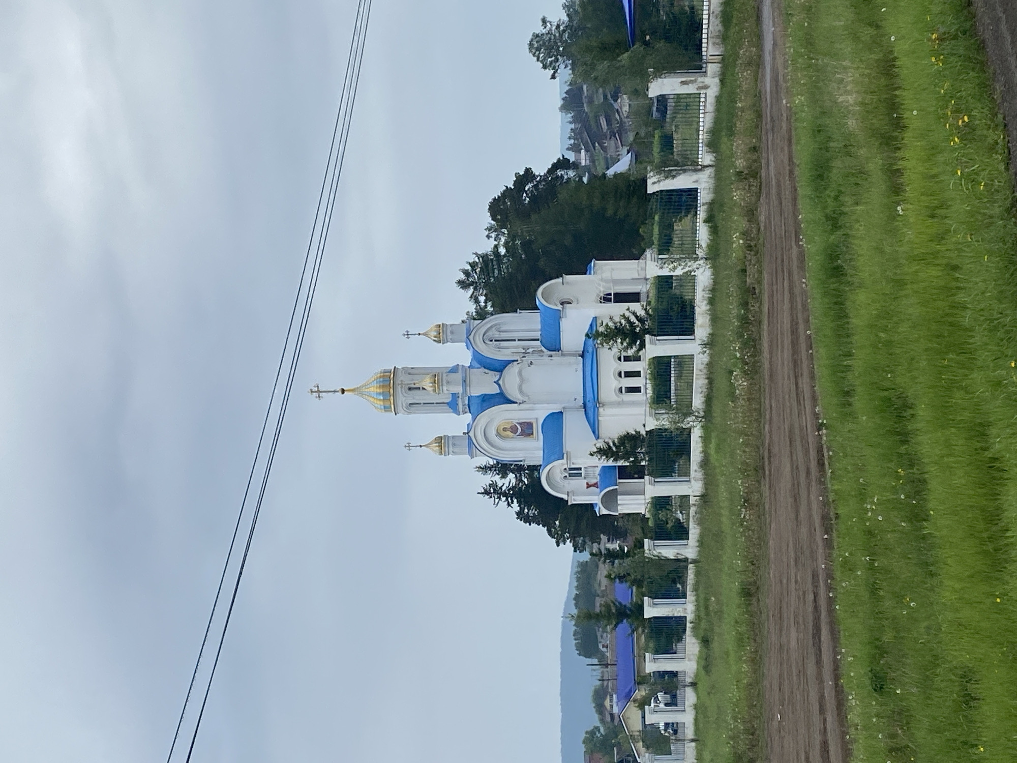 A white Orthodox church with blue domes and golden crosses stands on a hill, surrounded by green trees and a white fence. The church has several onion domes, the largest in the center topped with a golden cross and flag. In front of the church is a large grassy field and a dirt road. In the background, there are houses and hills. The sky is overcast. The location is the city of Pivovarikha, Russia.