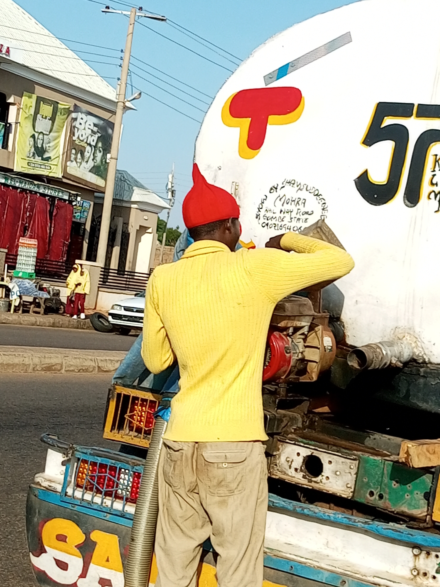 In Garko, Nigeria, a man wearing a yellow sweater and a red hat is working on a large white tank vehicle. The tank has a red and yellow graphic of the letter "T" and the number "5T" painted on its side. Below the graphic, there is handwritten text that appears to be contact information. The man is positioned behind the tank, near a red engine, and appears to be connecting a flexible pipe. The vehicle is parked on a paved road, with buildings and banners visible in the background. Another person in a yellow and red outfit is standing near a white car further down the road. The scene suggests an ongoing operation related to the water tank vehicle.