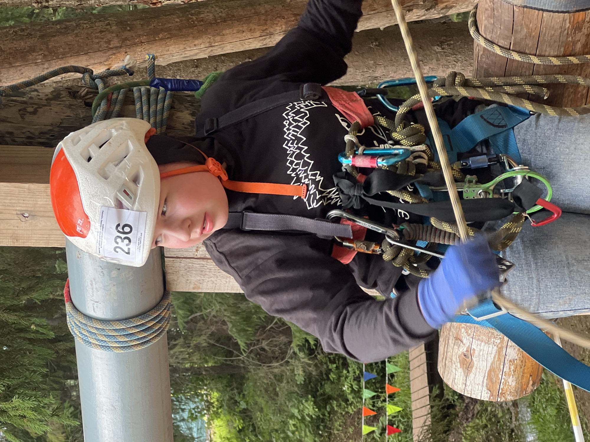 The image shows a person wearing a helmet and harness, suspended by ropes in what appears to be an outdoor adventure or obstacle course. The subject is wearing a white helmet with orange accents and a bib with the number "236" visible. They are also wearing blue gloves and a black hoodie with a white graphic design. The scene is set in a wooded area with trees and foliage visible in the background. Several ropes and carabiners are attached to the harness, indicating preparation for or participation in an activity like rock climbing or zip-lining. There are wooden structures, likely part of the course, and a series of colorful triangular flags in the background. The lighting suggests it might be daytime. The subject's expression is focused. The location context provided is Сергино, Russia.