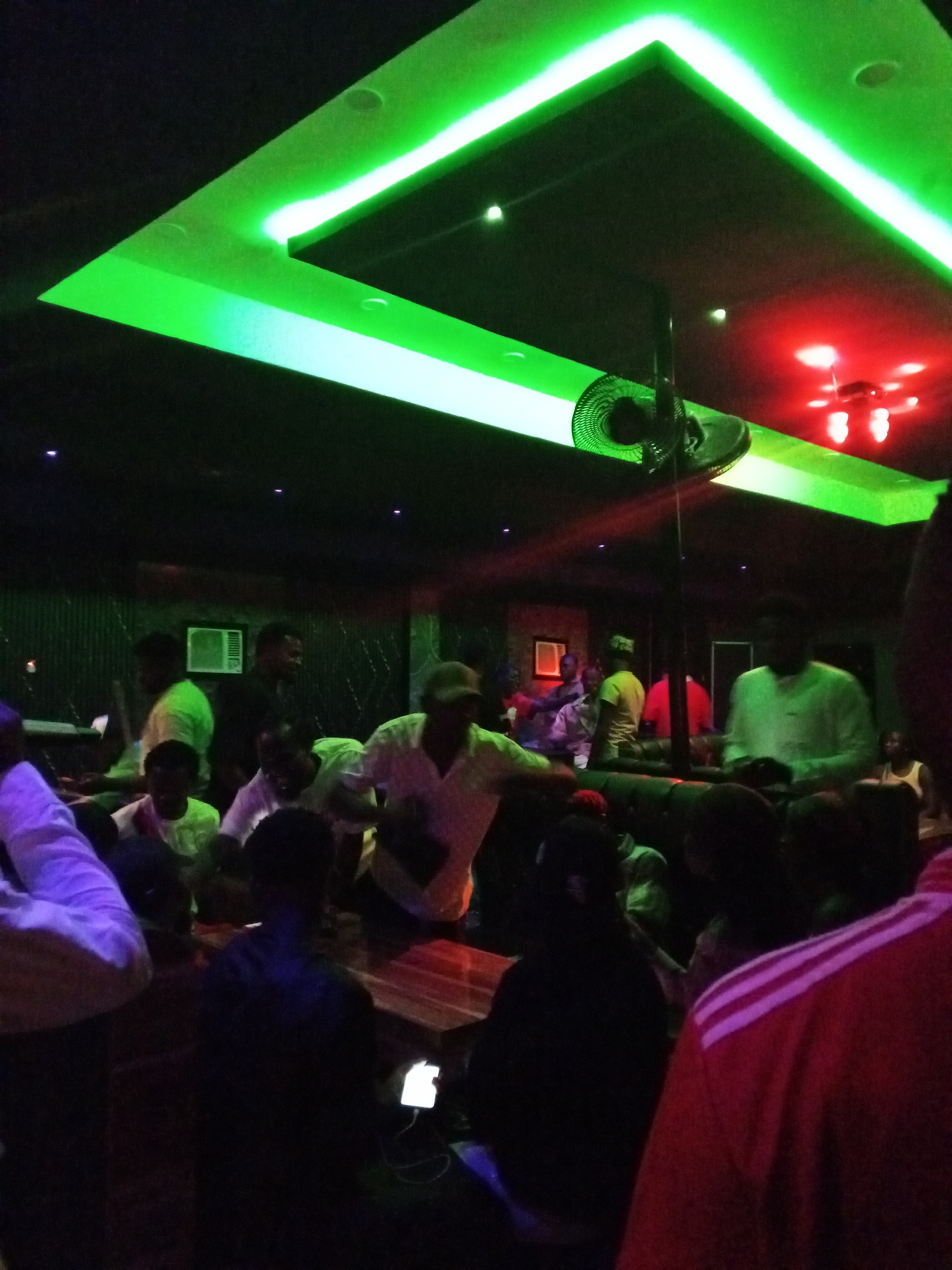 The image depicts a dimly lit interior space, likely a bar or club in Lokoja, Nigeria. A group of people are gathered around tables, some seated and some standing. The lighting consists of green neon strips outlining a square or rectangular section of the ceiling, along with red lighting in another area. A fan is visible hanging from the ceiling. Individuals are dressed in casual clothing. The overall atmosphere is one of a social gathering in an entertainment setting.