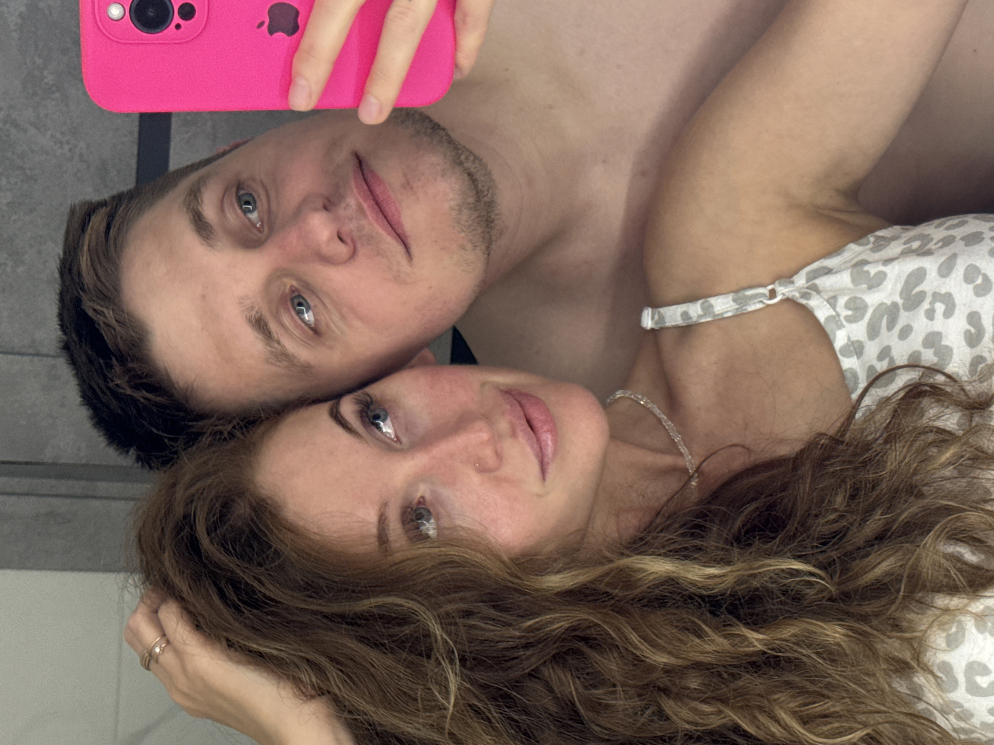A man and a woman are taking a selfie. The man is on top and is holding a pink cell phone. The woman is on the bottom, and she is wearing a white and gray top with thin straps. The background is a gray wall. The image may have been taken in an apartment or home. It is not possible to confirm the location is in Chaikovsky, Russia, from the image.