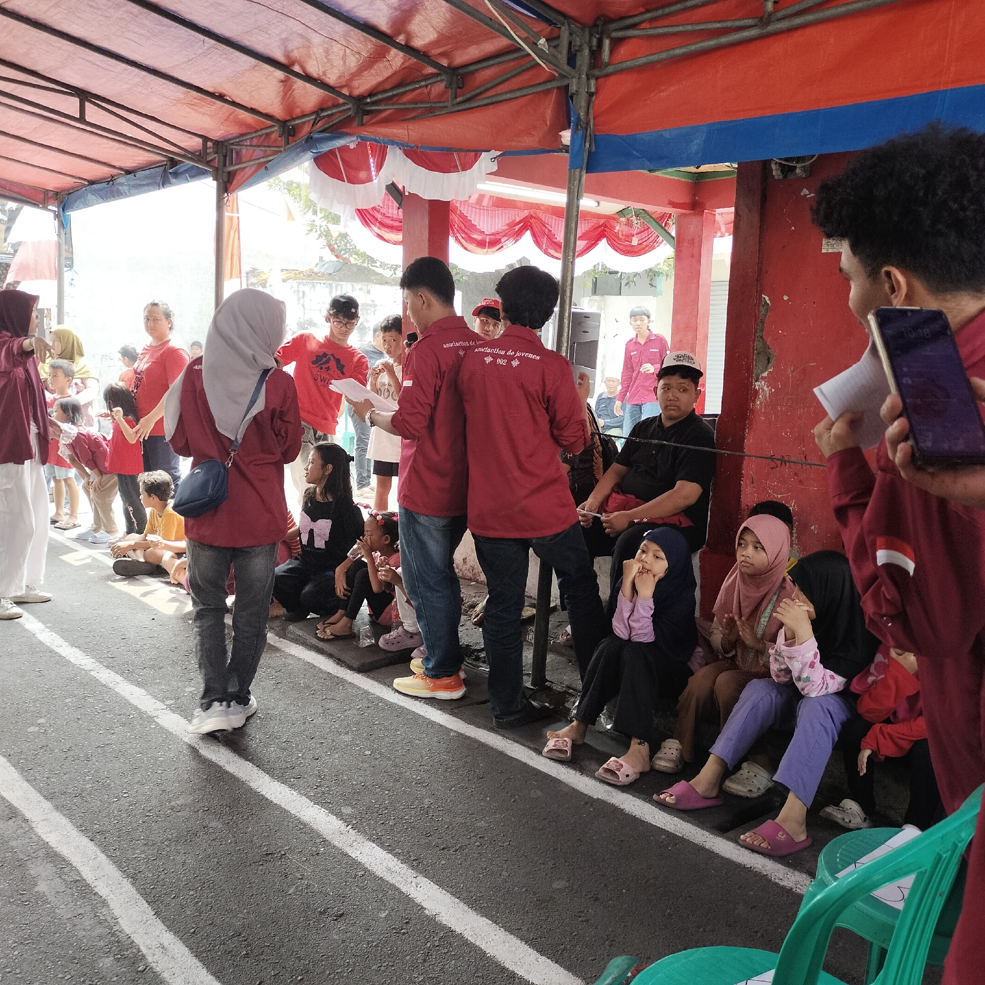 The image depicts an outdoor gathering under a red and blue tarp, possibly a community event or competition, likely located in Tangerang, Indonesia, given the presence of Indonesian flags and red and white decorations. A diverse group of people, including adults and children, are seen standing and sitting, some in maroon t-shirts with identical lettering on the back. The ground is asphalt with white lines, suggesting a designated area for activities. People are engaged in various activities, such as observing, participating, and socializing. The overall atmosphere appears casual and festive, with a focus on a communal event.