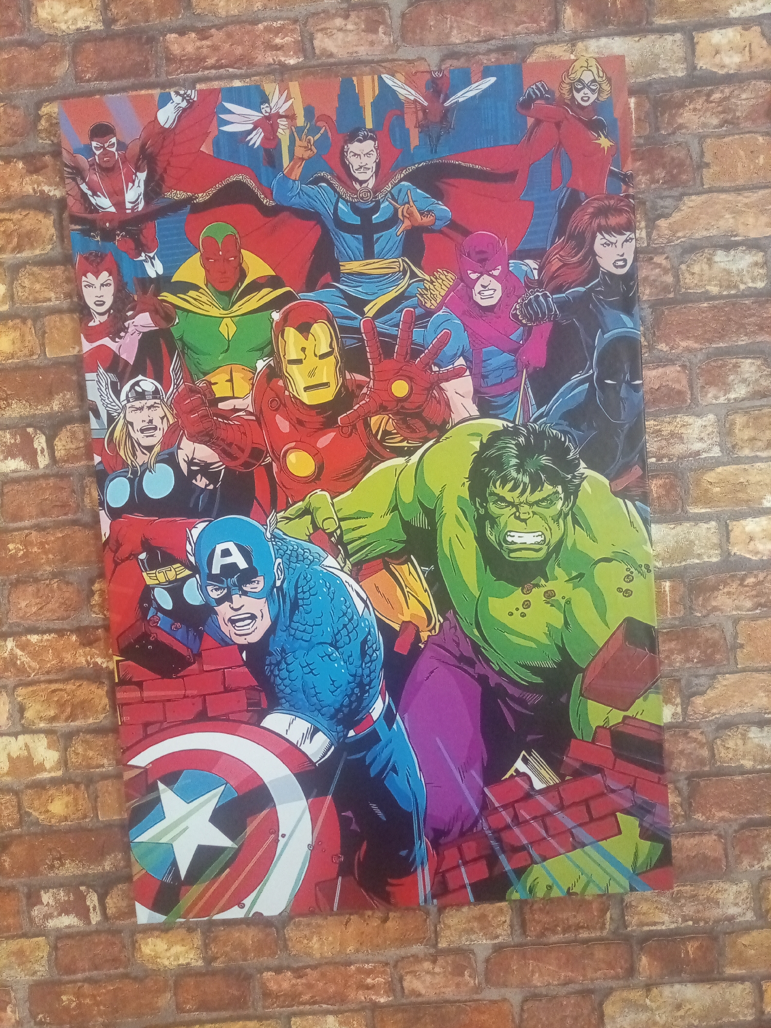 The image shows a rectangular Avengers poster depicting a variety of Marvel superheroes, including Captain America, Hulk, Iron Man, Thor, Vision, Doctor Strange, Black Widow, Black Panther, Captain Marvel, Falcon, Hawkeye, and Scarlet Witch, among others. The poster is affixed to a brick wall. There is no visual information about the location in the city of Porlamar, Venezuela.