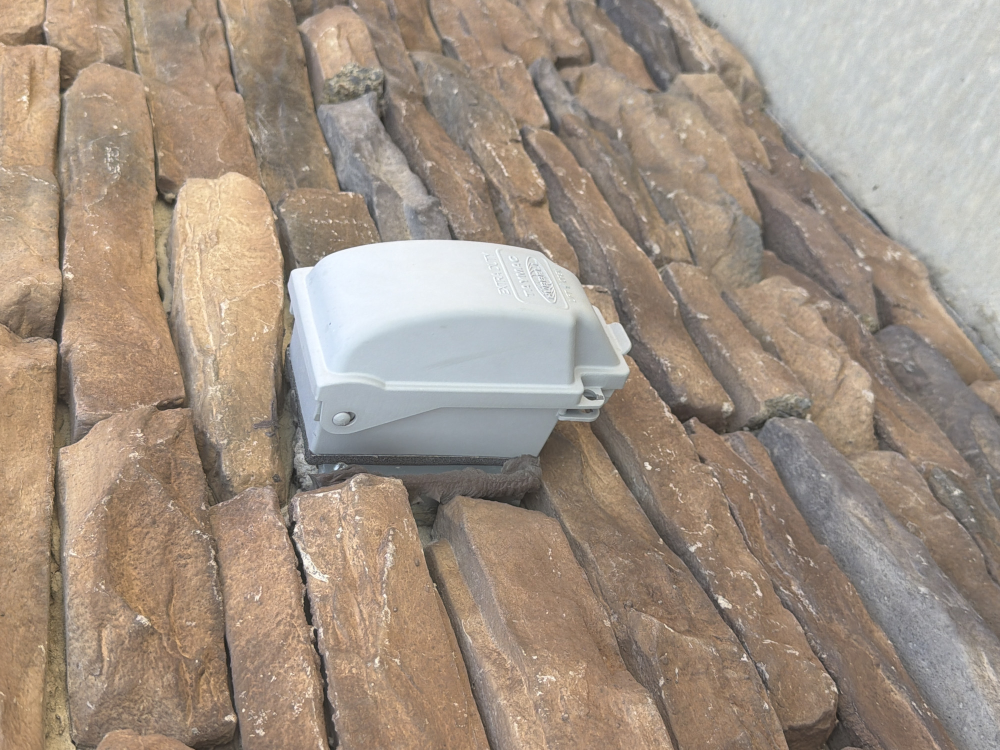 A light gray, rectangular electrical outlet cover is mounted on a stone wall. The stone is a mix of brown and gray hues, with varied shapes and sizes that create a textured, layered appearance. The cover has a circular button or latch visible on its top left. Some text is embossed on the cover, including "EXTRA-DUTY" and "TAYMAC". The outlet cover appears to be facing towards the viewer. The image is an outdoor, eye-level shot, with a shallow depth of field that blurs the background stones slightly. There are no people or animals visible in the image. The context of Cardwell, United States is unknown from the image alone.