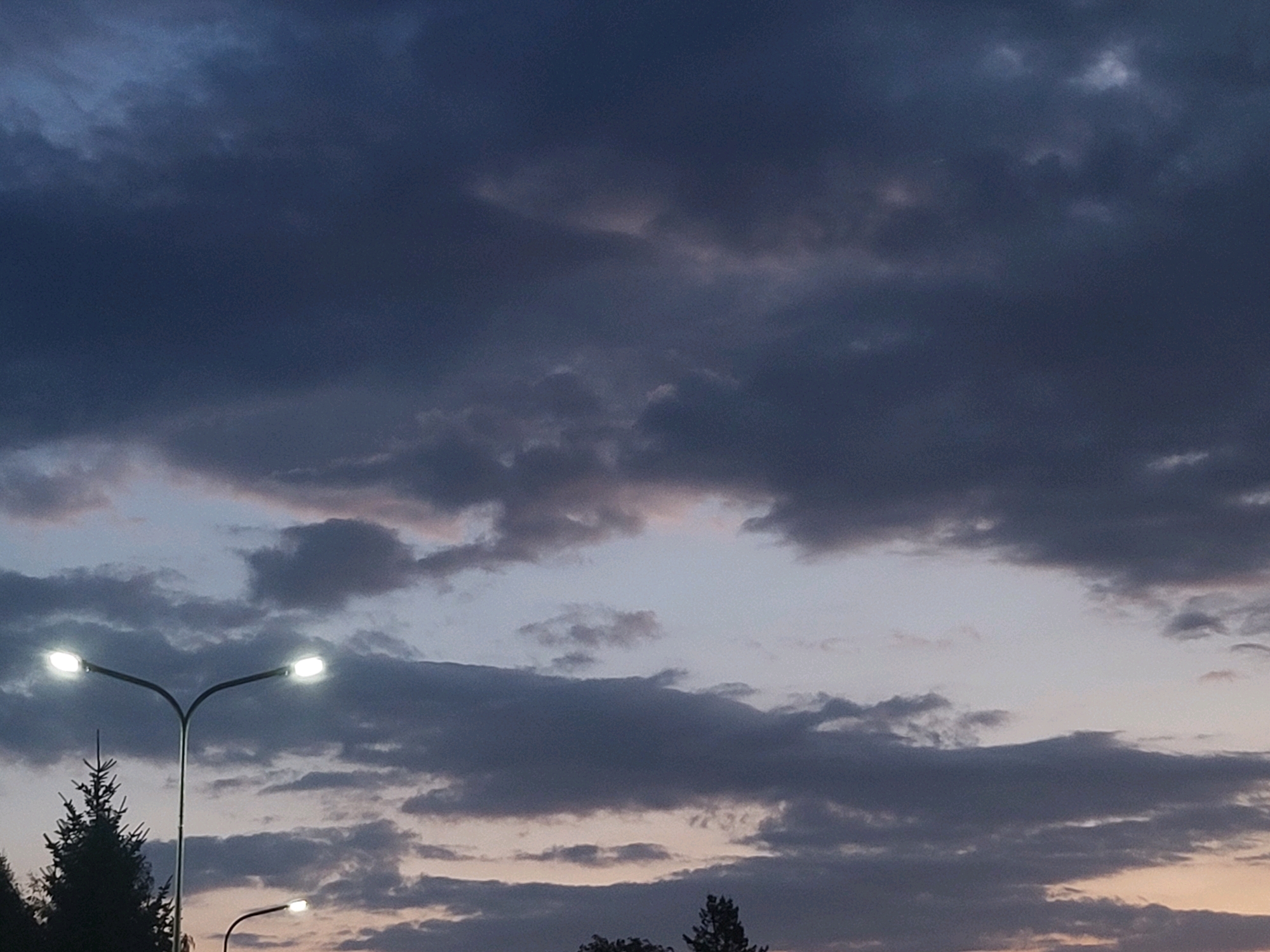 The image depicts a dusk sky over Ozolnieki, Latvia, characterized by a mix of dark, heavy clouds and lighter, wispy formations. Two streetlights are visible in the foreground. The larger streetlight, on the left, is a double-headed lamp with both lights illuminated, casting a bright glow. To the right and slightly lower, a single-headed streetlight is also lit. A silhouetted evergreen tree stands to the left of the double-headed streetlight, and other trees are visible as dark shapes in the lower part of the image, suggesting a semi-rural or suburban setting. The overall atmosphere is subdued, with the illumination from the streetlights contrasting against the darkening sky.