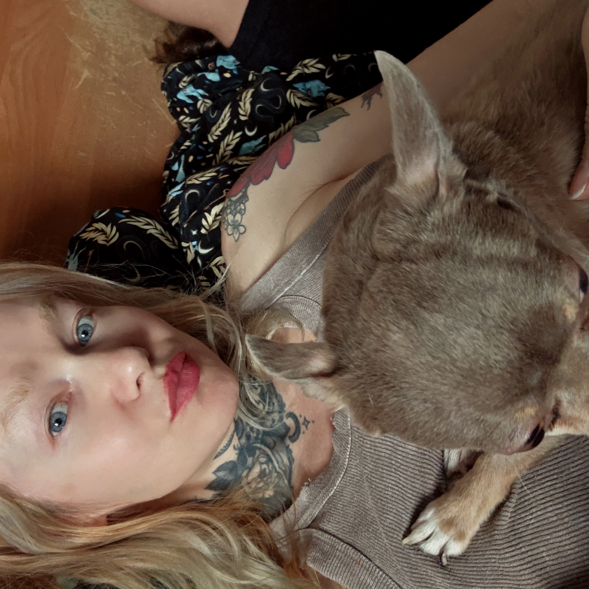 A woman with fair skin, blue eyes, and blonde hair is lying down, likely on a floor or bed, with a small brown dog resting on her chest. The woman is wearing red lipstick. She has multiple tattoos visible on her neck, chest, and shoulder. She is wearing a striped top and a black shirt with a pattern is nearby. The dog appears to be a chihuahua or similar breed. The lighting is somewhat dim and possibly indoor. There is no information to indicate that this image was taken in Saint Petersburg, Russia.