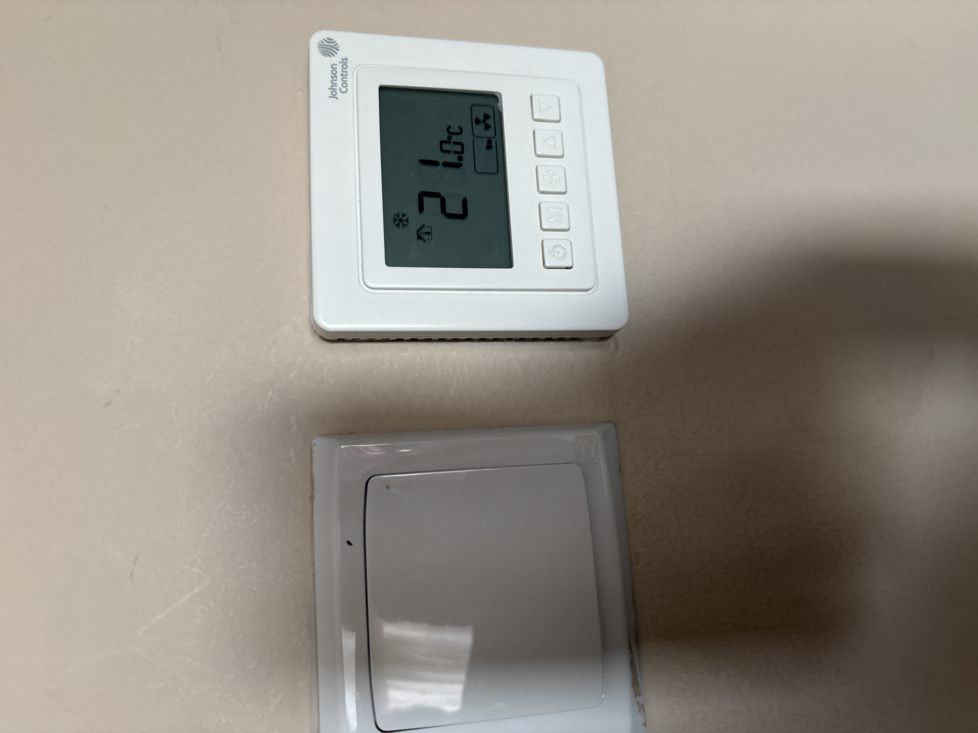 The image shows a close-up of a white thermostat with a digital display and several buttons on a light-colored wall. The thermostat displays a temperature of 21.0°C, a house icon, a snowflake icon, and a fan icon. The brand name "Johnson Controls" is visible at the top of the thermostat. To the left of the thermostat is a white light switch. The wall appears to be painted and has some faint marks on its surface. There is no indication of the location being Dubai, United Arab Emirates.