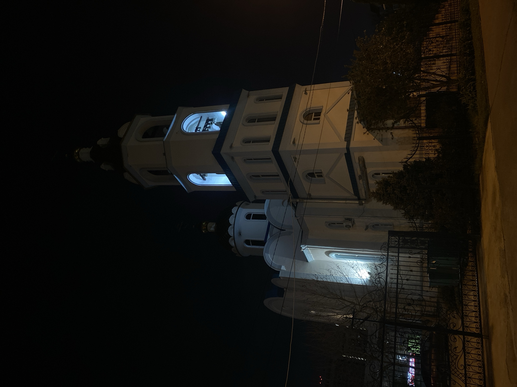 Captured at night, the image showcases a church in an unknown urban setting. The church, illuminated, stands against a dark sky. Its architectural features include a tall bell tower with a blue-lit interior, arched windows, and a white facade accented with darker trim. A wrought-iron fence borders the church, with trees and foliage partially obscuring the view. The environment is dim, with artificial lights highlighting the church's structure.