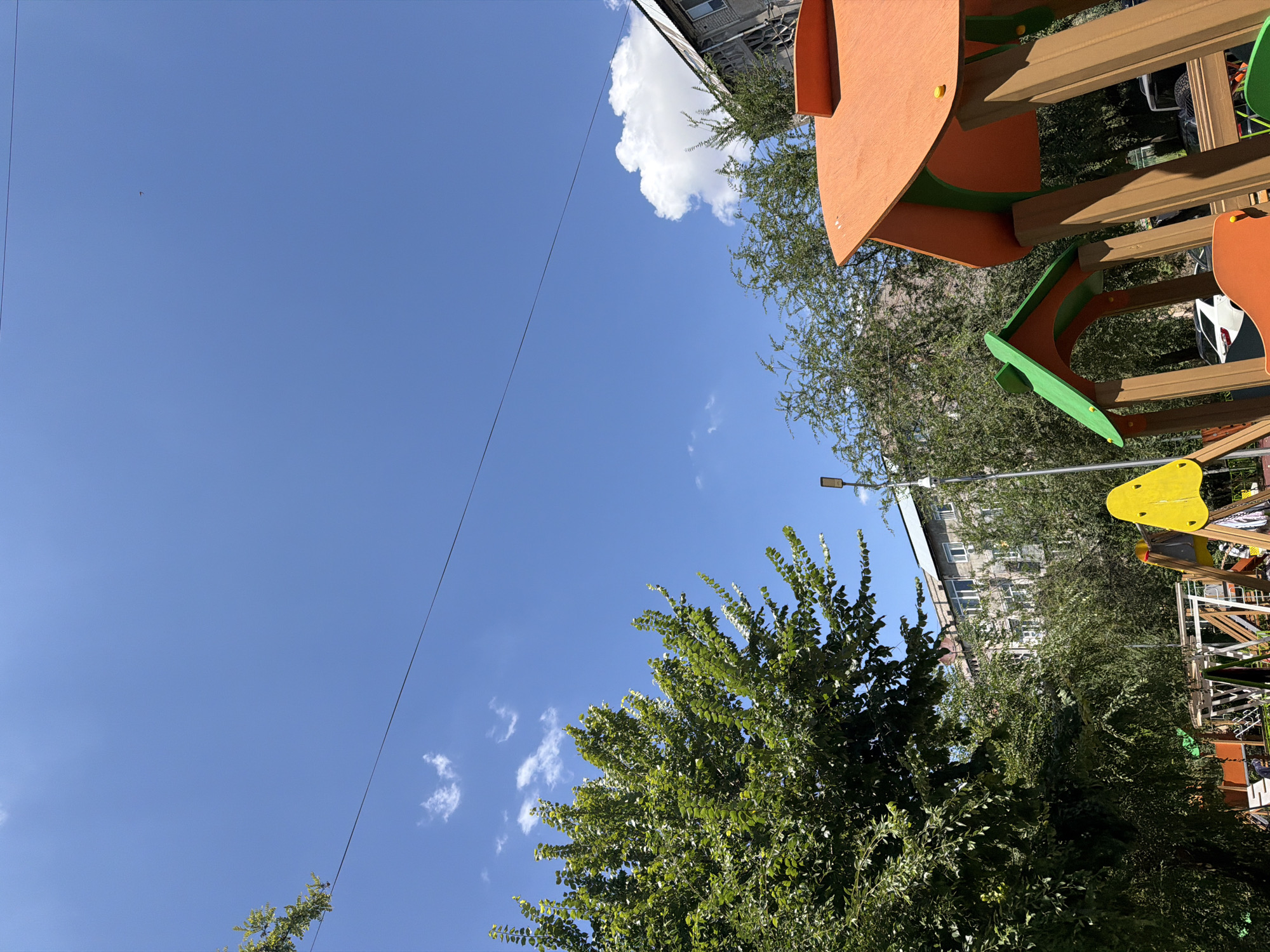 The image depicts a children's playground with colorful structures under a clear blue sky. A large, leafy green tree dominates the left side of the frame, with its branches extending upwards. In the foreground, parts of a playground set are visible, including an orange roofed structure and a green arched roof. A bright yellow slide or climbing element is also present. Behind the playground, several apartment buildings are visible, suggesting an urban setting. The sky is clear with a few scattered white clouds. A thin black wire stretches horizontally across the upper portion of the image. The presence of apartment buildings and the style of the playground suggest a residential area within the city of Shymkent, Kazakhstan. There are no people explicitly visible in the frame.
