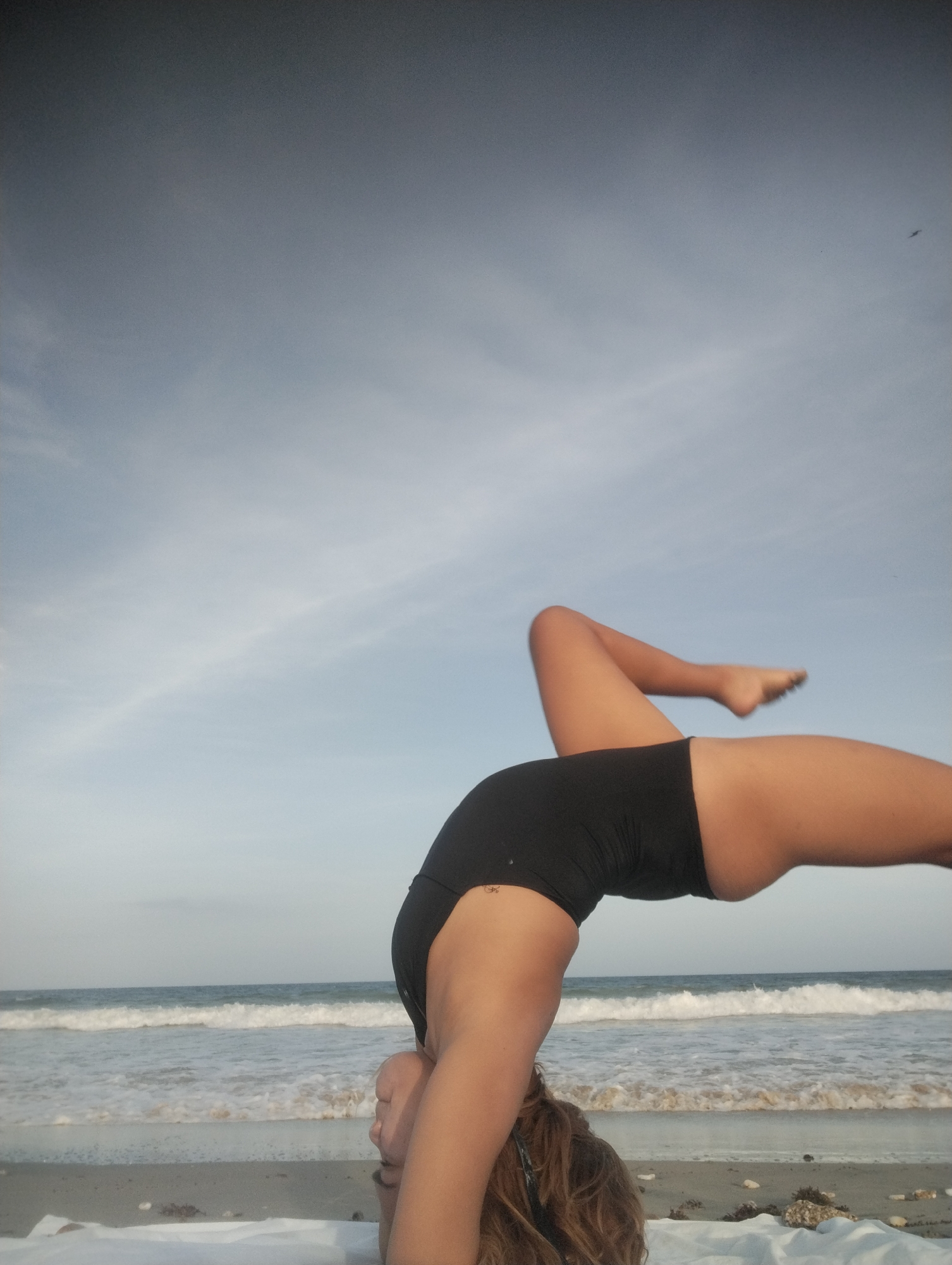 A person is performing a yoga pose on a beach. The person is in a headstand position, with their body arched backward. They are wearing a black swimsuit. The beach has light-colored sand, and there are waves visible in the background. The sky is partly cloudy. It is not possible to confirm that the location is the city of Porlamar, Venezuela.