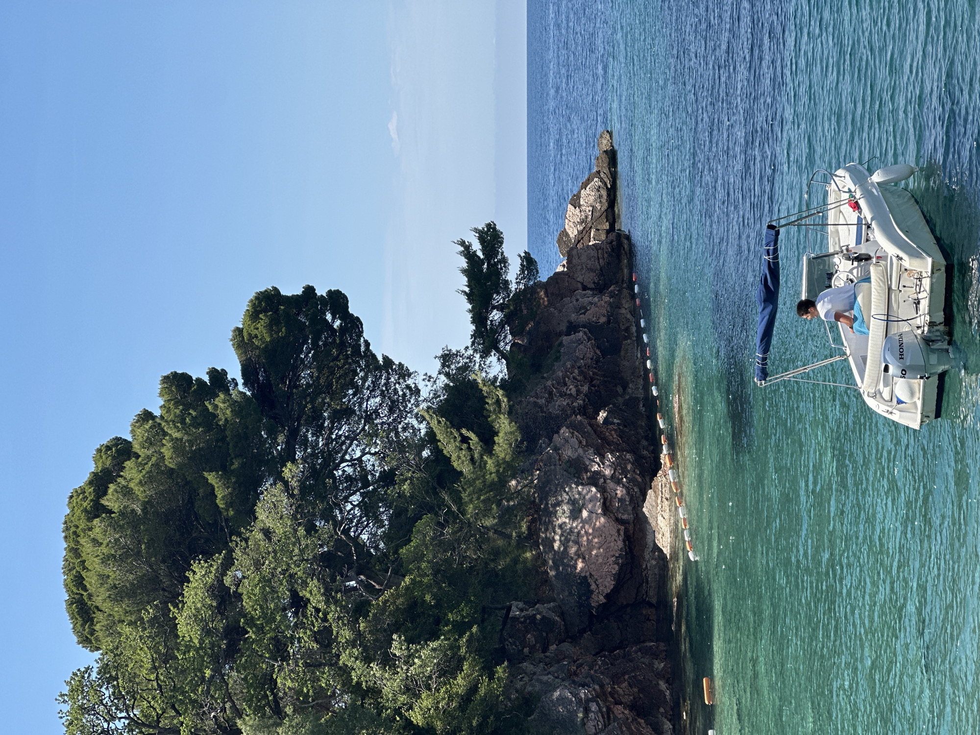 The scene depicts a rocky coastline in Sveti Stefan, Montenegro, adjacent to clear turquoise waters. Lush green trees cling to the rocky outcrop. A small white sailboat is anchored near the rocks, with a man aboard, appearing to tend to the boat. Buoys line the water's edge, likely marking a boundary. The sky above is clear and blue, indicating a sunny day.