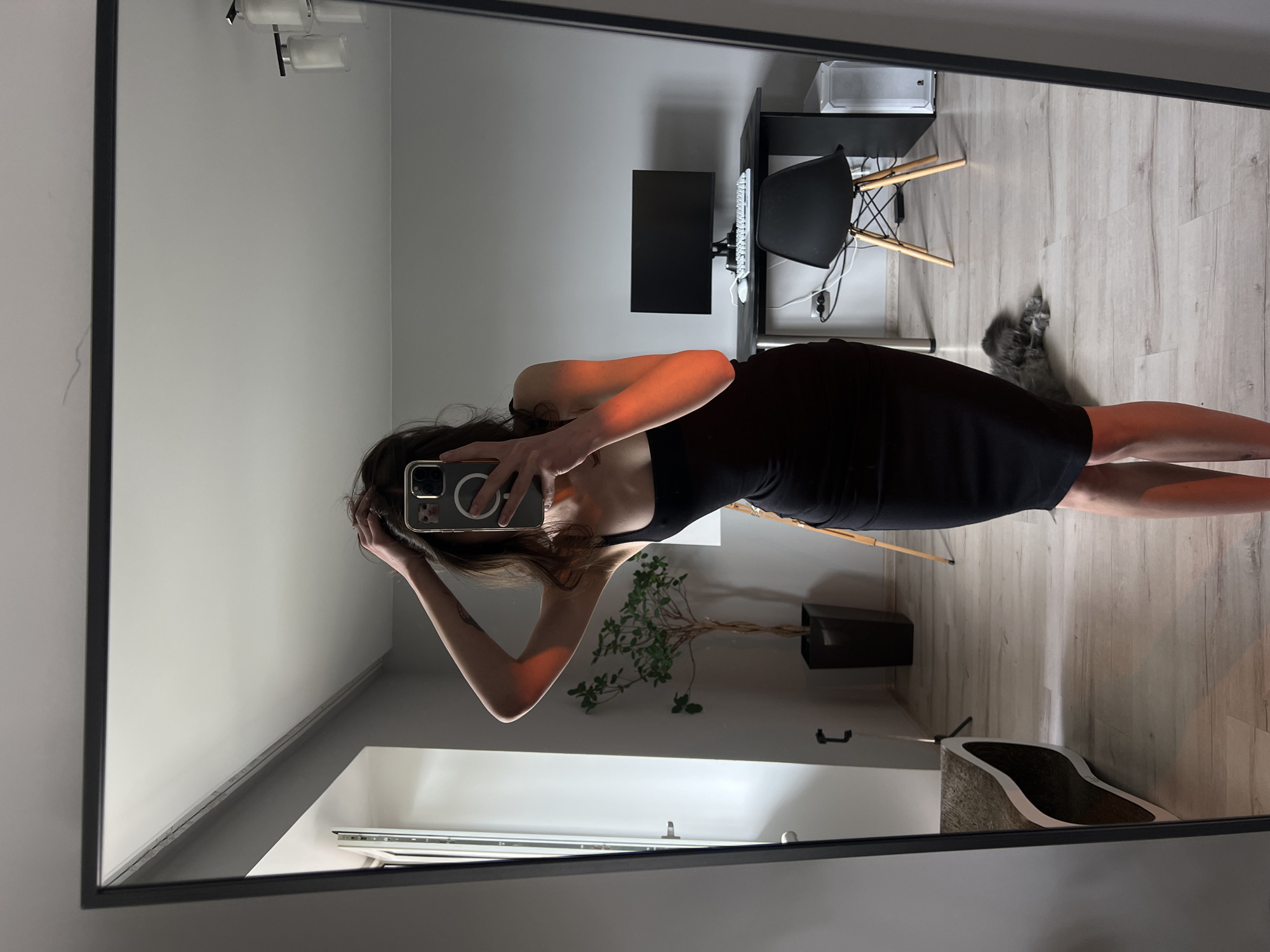 In a room with light gray walls and a light-colored laminate floor, a young woman with dark hair is taking a mirror selfie. She is wearing a black, form-fitting, knee-length dress and has her right hand in her hair. An orange light casts a shadow on her right arm and shoulder. In the reflection, a black desk with a computer monitor, keyboard, and mouse is visible against the wall. A black chair with wooden legs is pulled up to the desk. To the left of the desk, a white computer tower sits on the floor. A tall, dark pot with a leafy green plant stands to the left of the desk. In the lower right portion of the mirror's reflection, a long-haired gray cat is lying on the floor. To the left, a modern-style cat scratching post with a curved design is partially visible. A large, rectangular mirror with a thin black frame dominates the foreground.