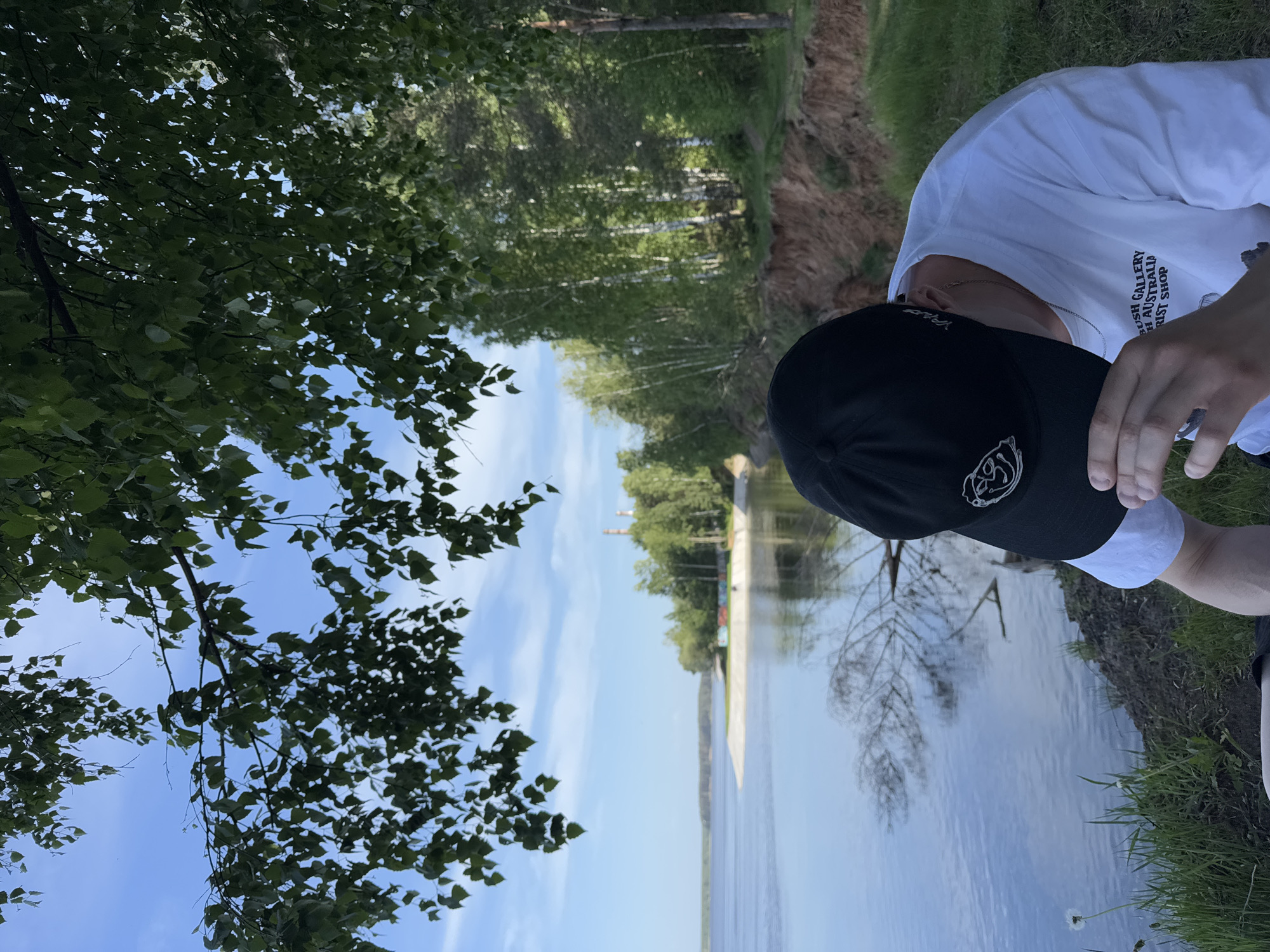 A person wearing a black baseball cap and a white t-shirt stands at the edge of a body of water, likely a lake or river. The environment includes trees and vegetation typical of a natural area. The sky is blue with some clouds. The location is the city of Chaikovsky, Russia. In the background, there are what appear to be industrial structures or tall buildings beyond the trees, possibly related to the industrial activity in the city. The person is on a high river bank.