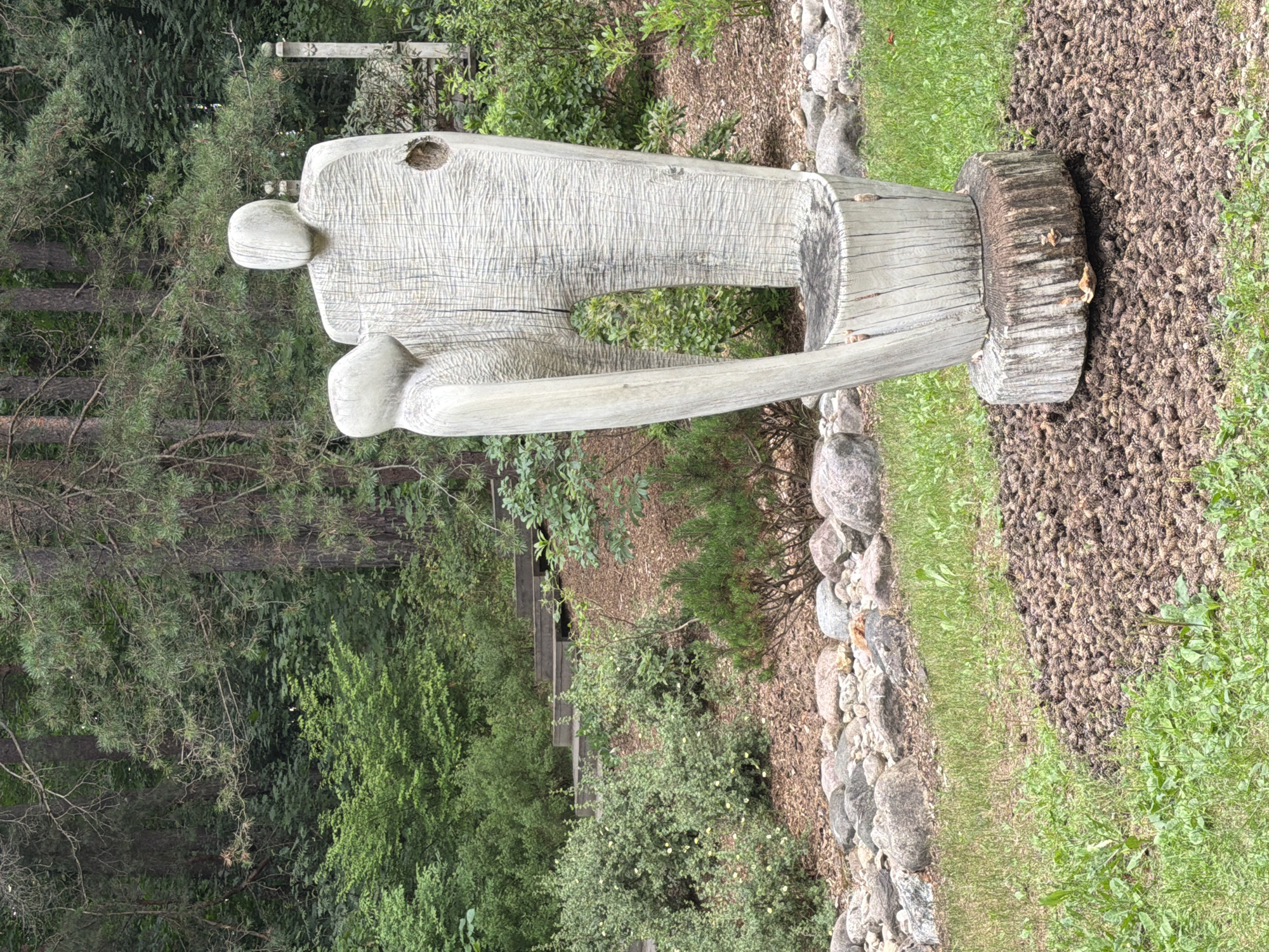 In Riga, Latvia, at an outdoor location characterized by dense forest and manicured landscaping, a weathered wooden sculpture of two abstract human figures stands prominently. The sculpture, with a rough, textured surface, depicts one figure taller than the other, positioned as if in conversation or embrace. The larger figure has a hollowed-out section on its right side. The sculpture is mounted on a thick, cylindrical log base, which is surrounded by a bed of pinecones and scattered green vegetation. In the background, a sloped area features a wooden staircase ascending through lush greenery, flanked by rocks and shrubs. The overall environment suggests a park or garden setting within a wooded area.