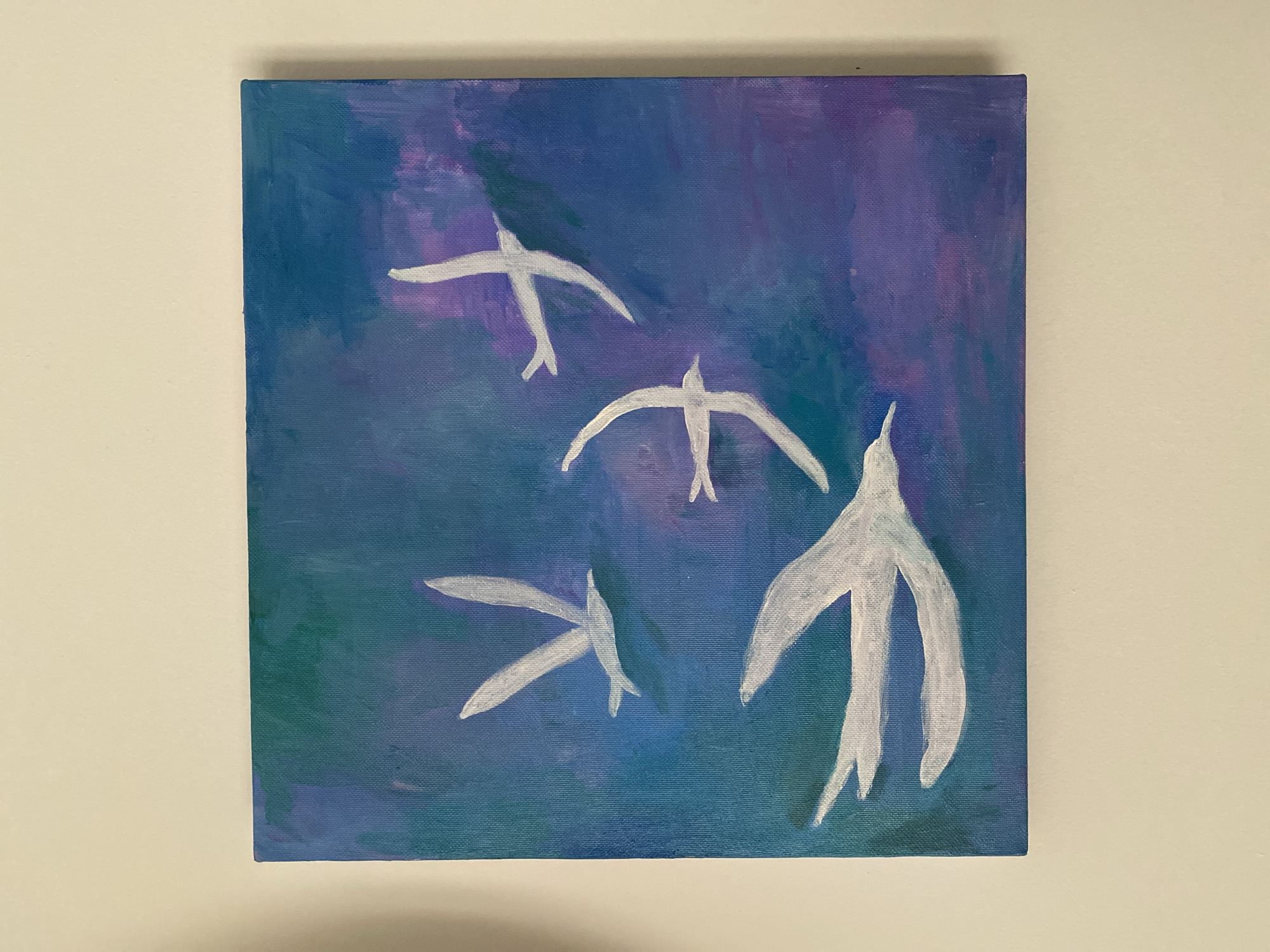 A square painting hangs against a neutral wall. The painting has a blue and purple background. Four white, stylized bird shapes are painted over the background. The shapes loosely resemble birds in flight. The painting's edges are visible.