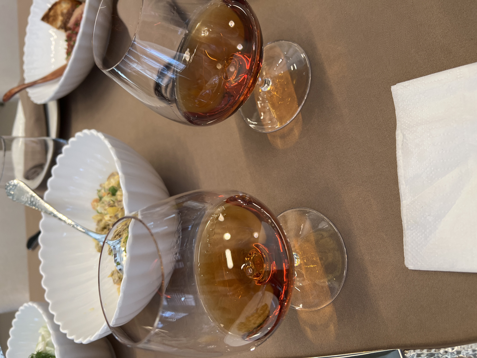 The image captures a table setting. Two partially filled wine glasses, containing a dark amber liquid, are positioned on the right side of the frame. A white linen napkin sits adjacent to the glasses. On the left side, there are three white, scalloped-edged bowls. Two bowls contain food, with the middle bowl including a silver fork. The table's surface is a textured brown. It is not possible to determine from this image if the location is in the city of Saint Petersburg, Russia.