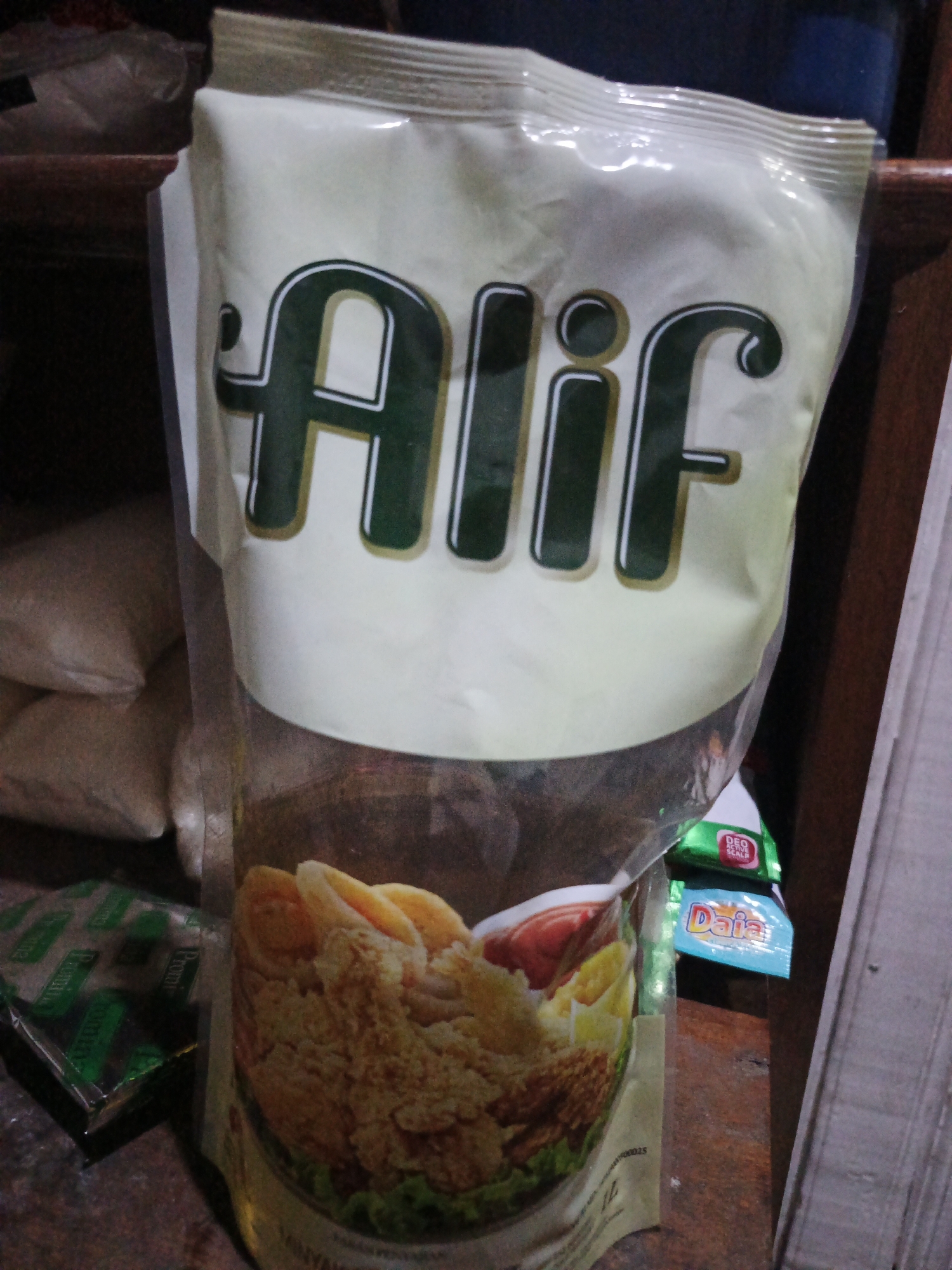 The image shows a pouch of cooking oil with the brand name "Alif" prominently displayed in large green lettering. The pouch has a picture of fried chicken on it. To the left of the Alif pouch, there are two smaller green packages, possibly sachets of seasoning or spices. To the right of the Alif pouch, there is a small blue packet with the word "Daia" on it, and above that, a green packet with red lettering. The background appears to be a wooden surface, possibly a shelf or table, and other indistinct items are visible in the background, including what looks like more bags or containers. The lighting suggests an indoor setting, likely a home or a small shop. There are no people visible in the image. The scene is not specific to Sinar Bulan, Indonesia, or any other particular city; it depicts common consumer goods.